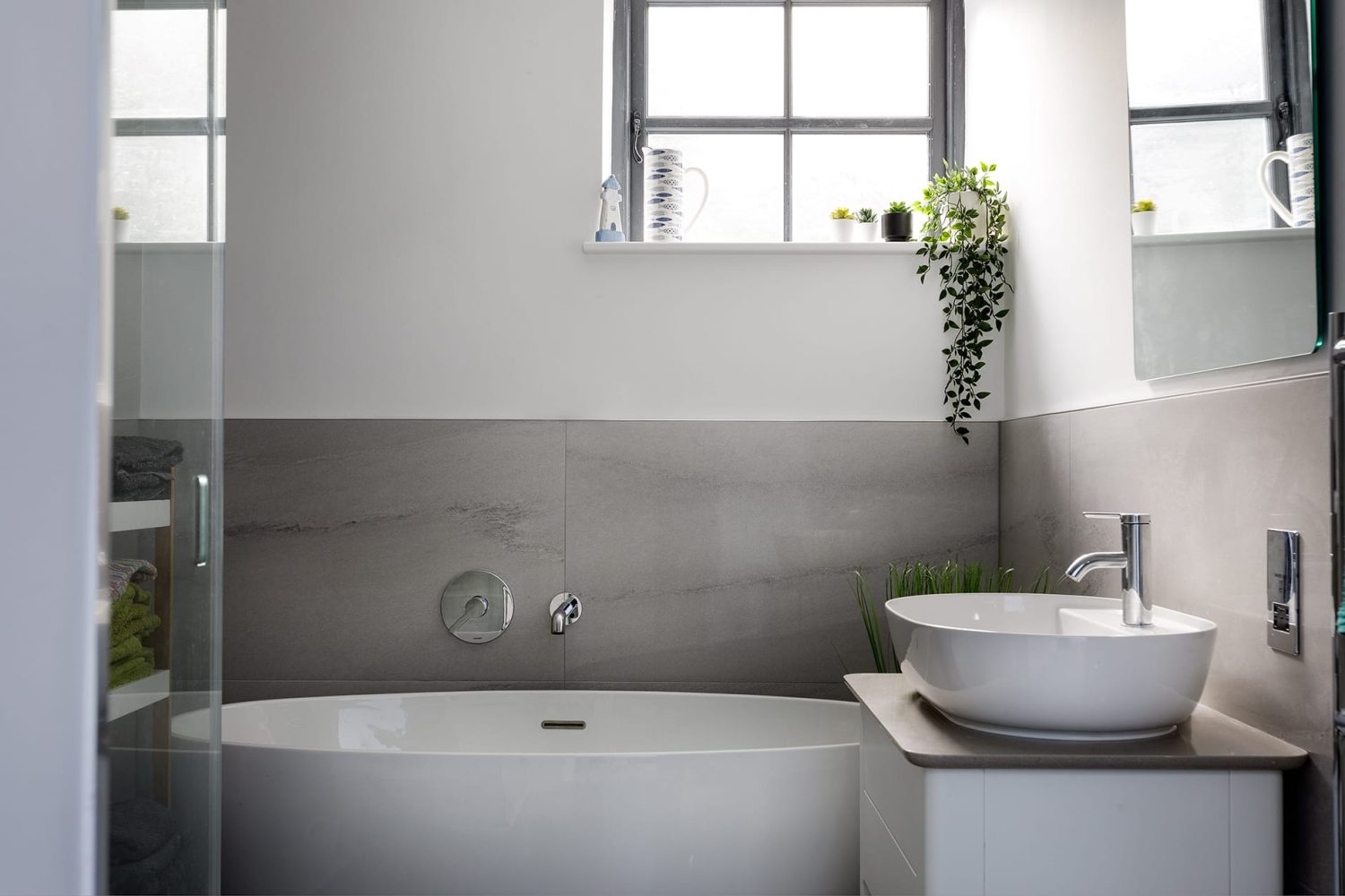Close up white sink with chrome tap in front of large white bath with grey tiled wall behind and plant hanging from windowsill