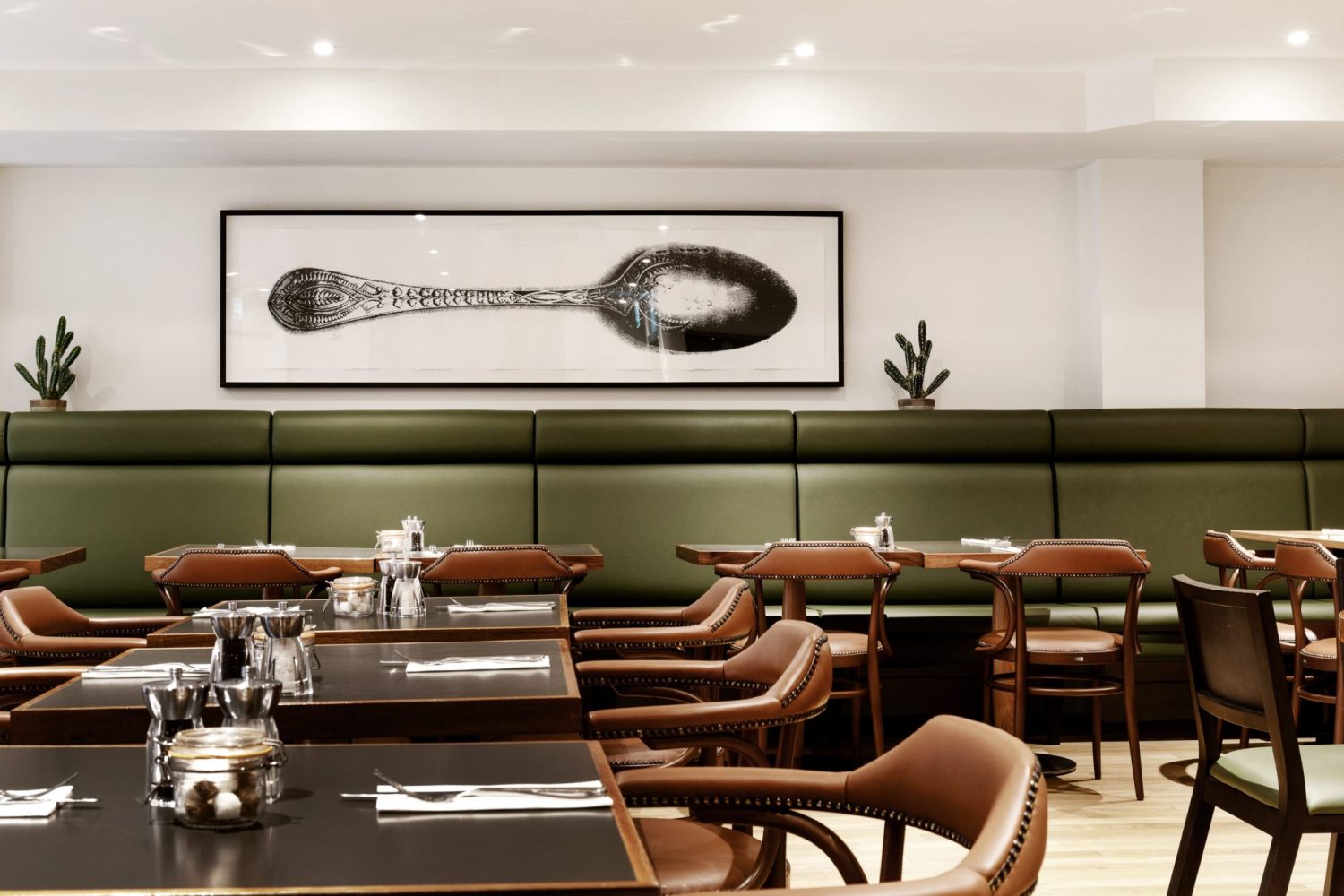 Cafe seating area with green leather booth seats across wall and black tables with brown leather chairs. Large framed image of a spoon on the wall