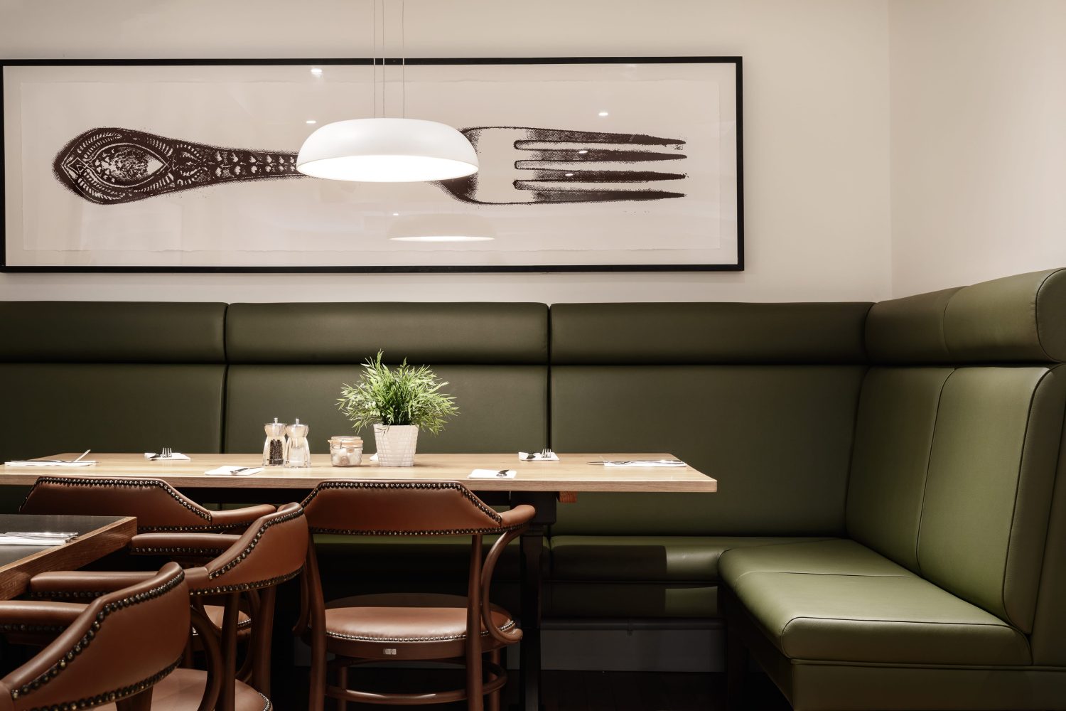 Cafe seating with a green leather corner booth seat with a wooden table and brown leather chairs. Large framed image of a fork on the wall above