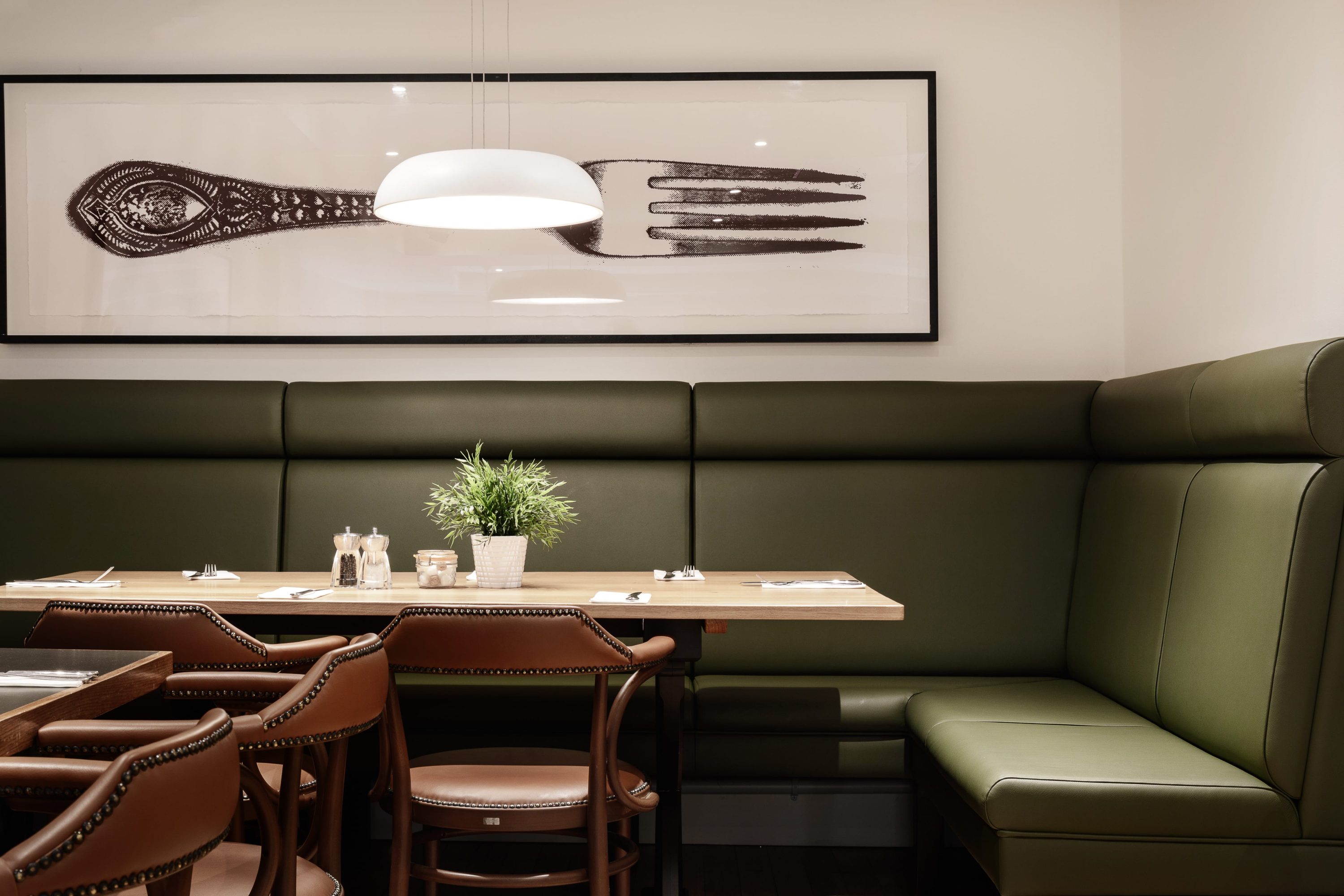 Cafe seating with a green leather corner booth seat with a wooden table and brown leather chairs. Large framed image of a fork on the wall above