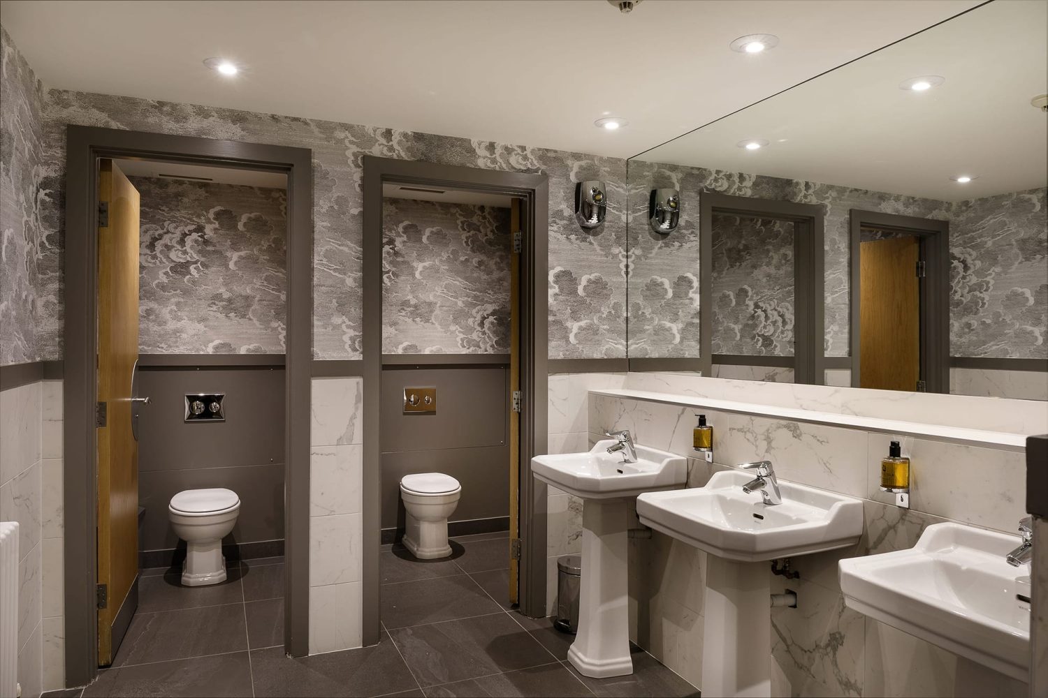 Bathroom with light grey wallpaper and white marble walls. Two toilet stalls and three white sinks with chrome taps and a large rectangular mirror above
