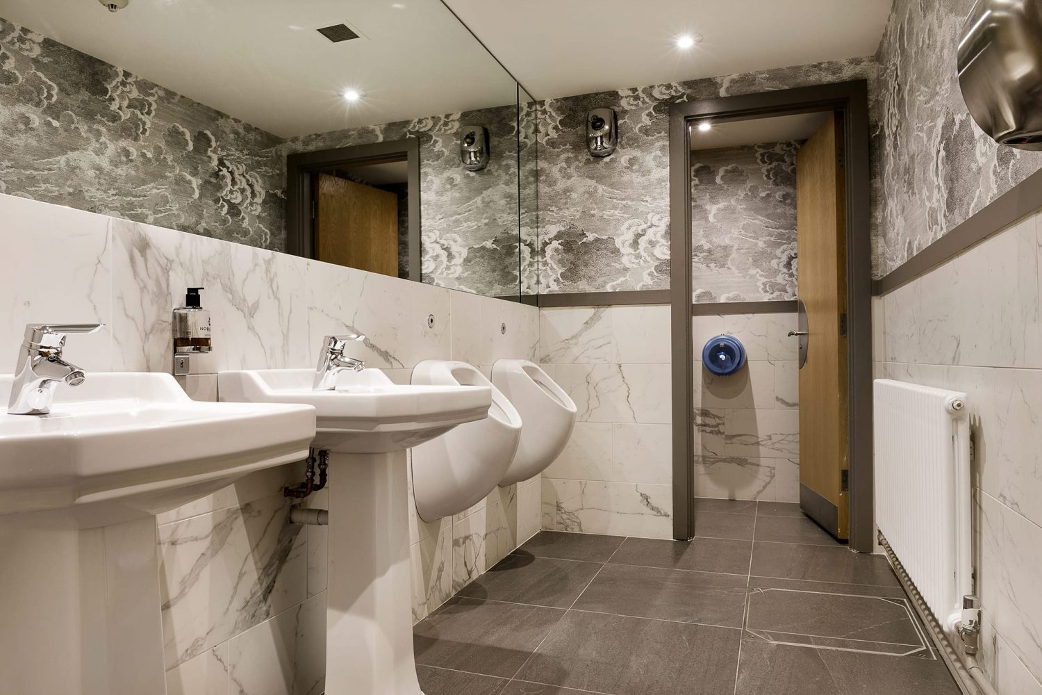 Bathroom with light grey wallpaper and white marble walls. One toilet stall and two white sinks with chrome taps and a large rectangular mirror above