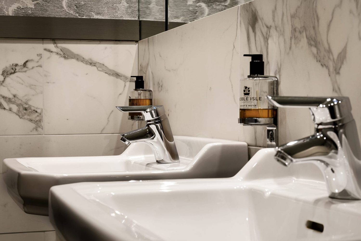 Two white bathroom sinks with chrome taps and soap holders attached to the white marble tiled wall
