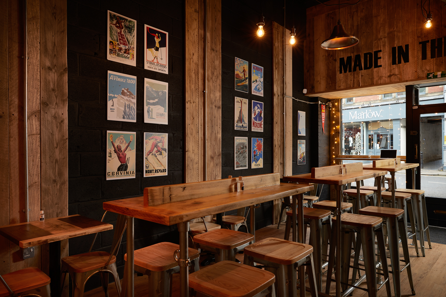 Cafe seating area with wooden tables and stools with black painted brick wall featuring ski-inspired posters
