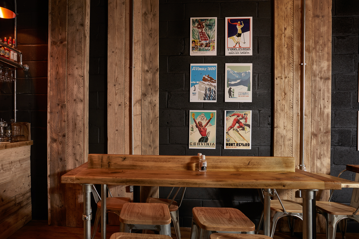 Cafe seating area with wooden table and stools with black painted brick wall behind featuring ski-inspired posters