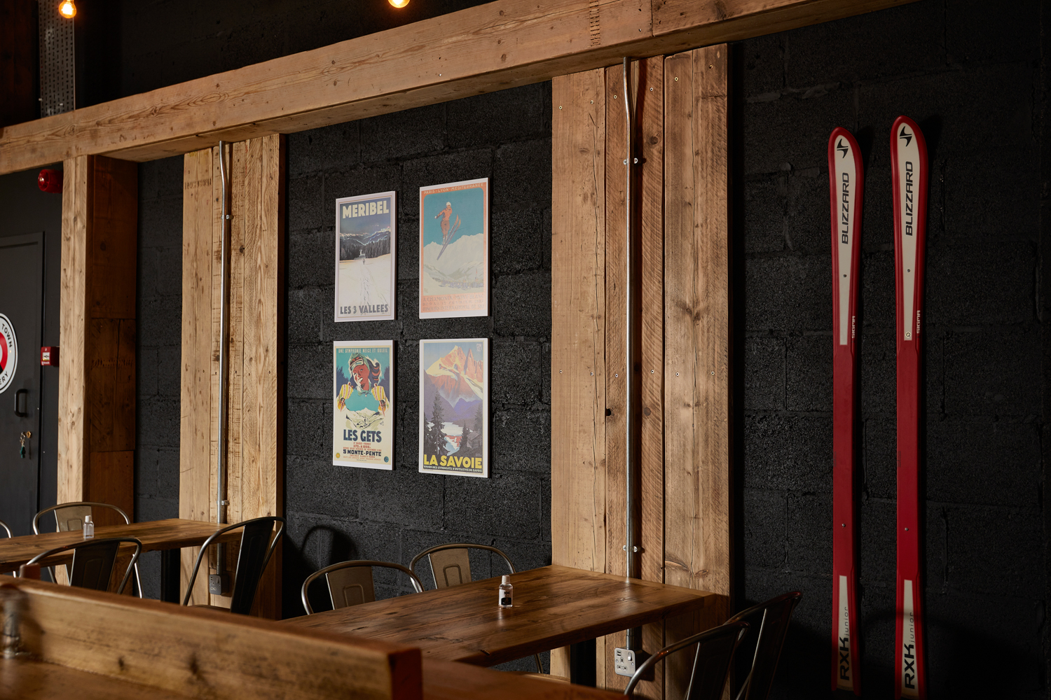 Cafe seating area with wooden tables and metal chairs with black painted brick walls featuring a pair of red skis and ski-inspired posters