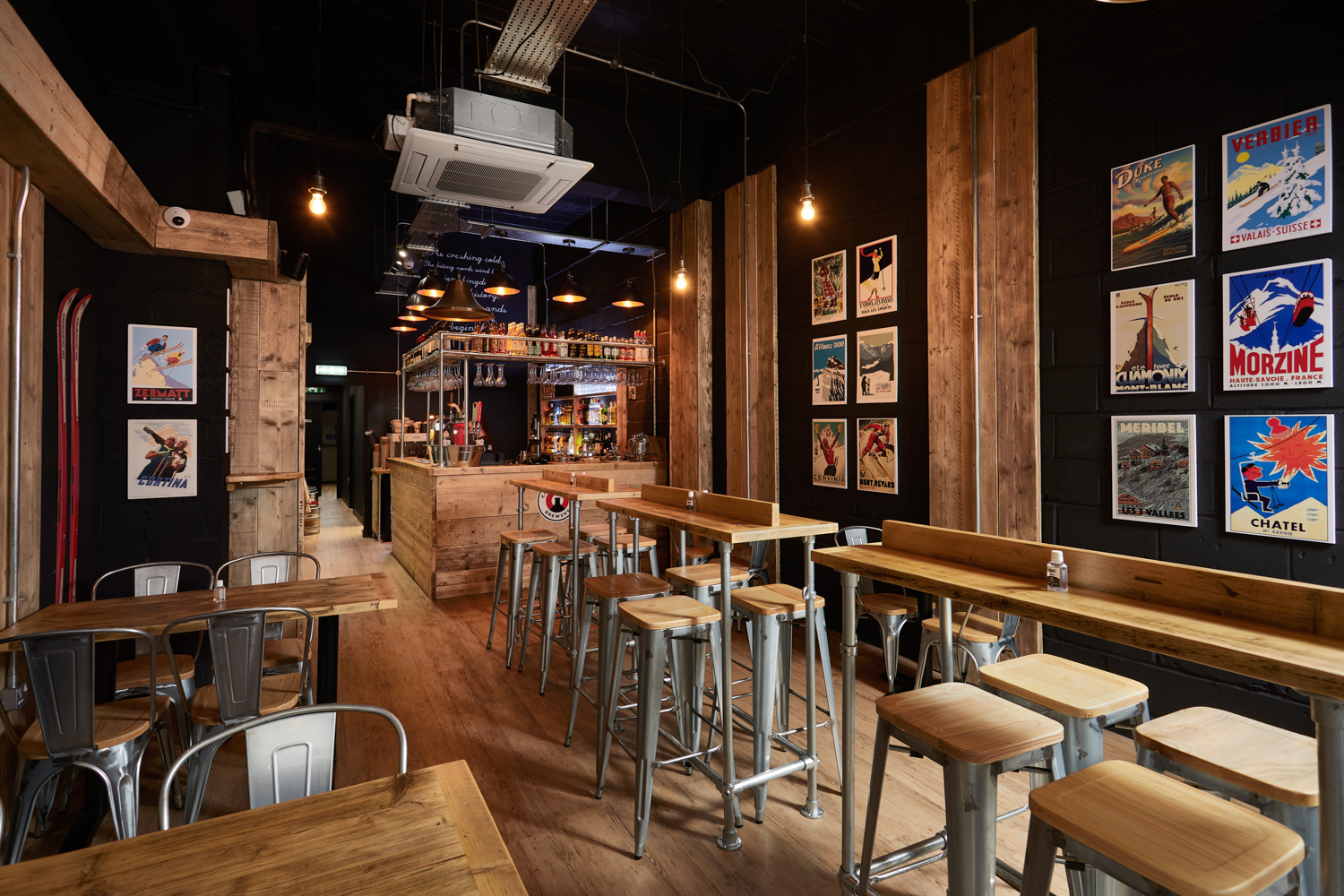 Cafe seating area with wooden tables and stools with ski-inspired posters on walls and bar area to the back