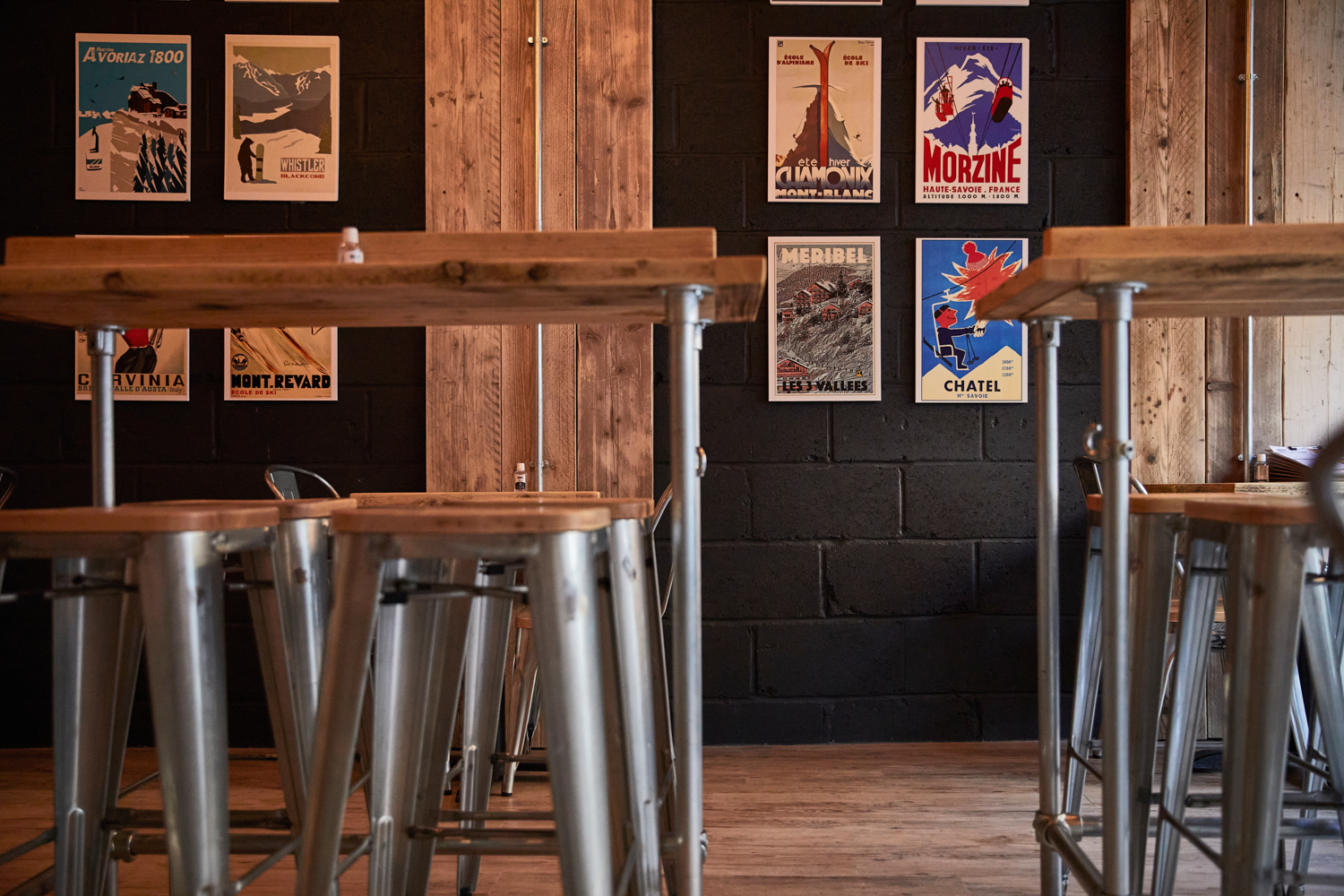 Cafe seating area with wooden tabletops and silver metal stools with black painted brick walls featuring ski-inspired posters