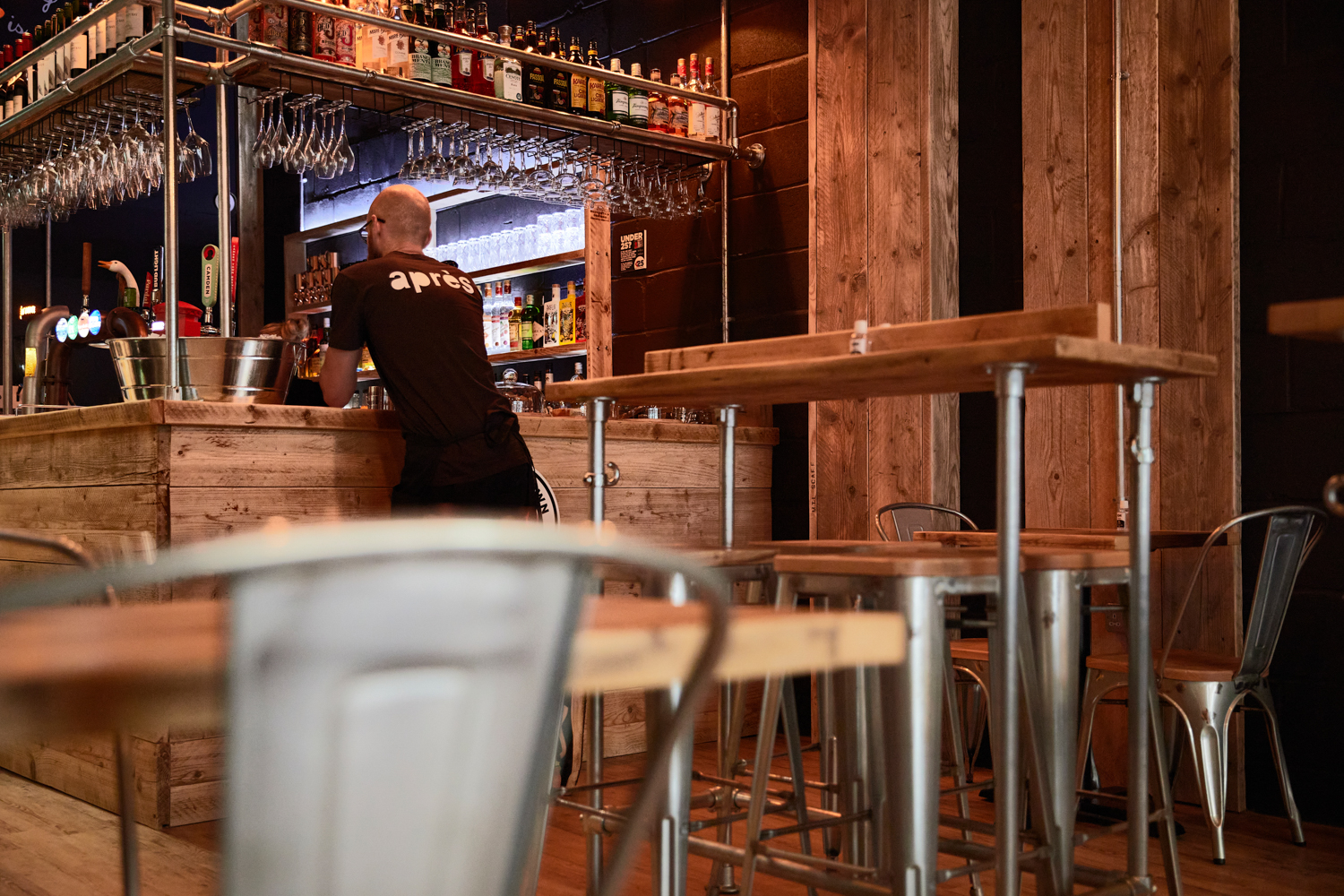 Man standing at bar with glasses hanging from shelving and wooden tables and seating behind him