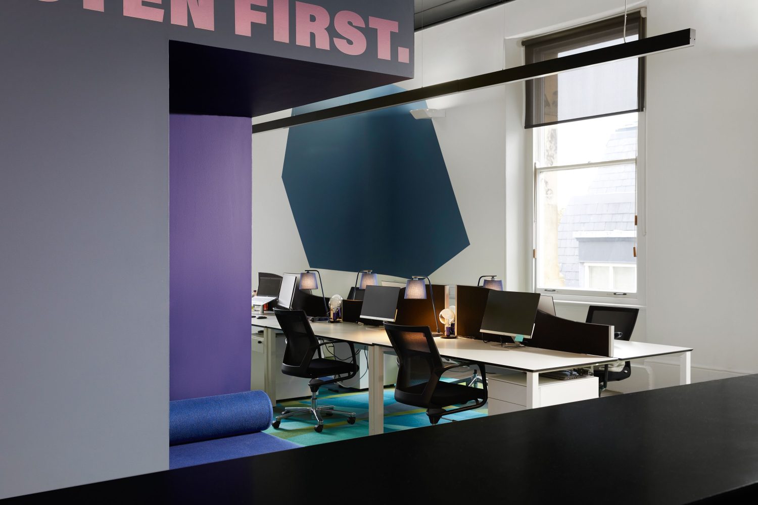 Open plan office desk space with a purple seating area build into the wall and a large blue geometric graphic on white wall