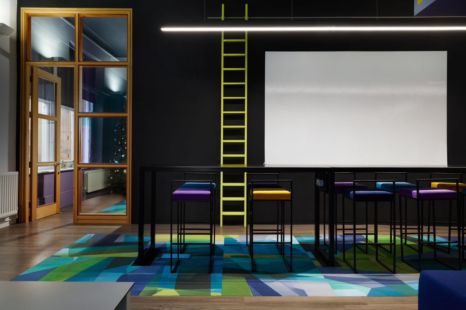 Long table with multi-coloured seats with a blue and green geometric floor beneath and whiteboard beside tall yellow ladder on the wall behind