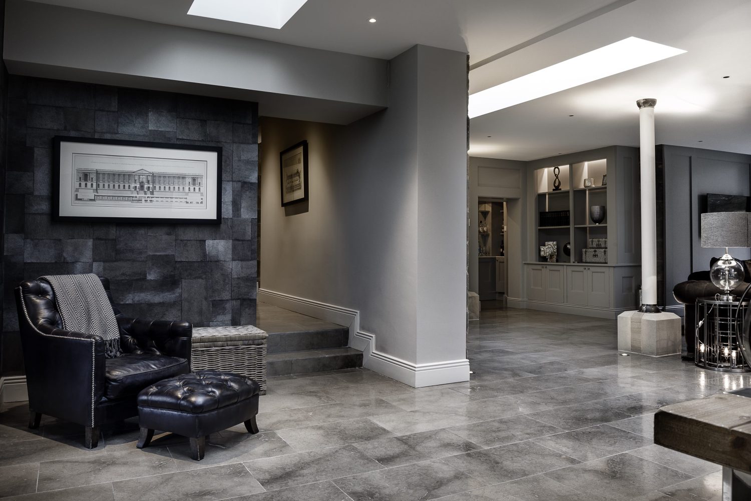Open plan living space with grey walls of different textures and grey tiled floor. Black leather seat with footstool to the left