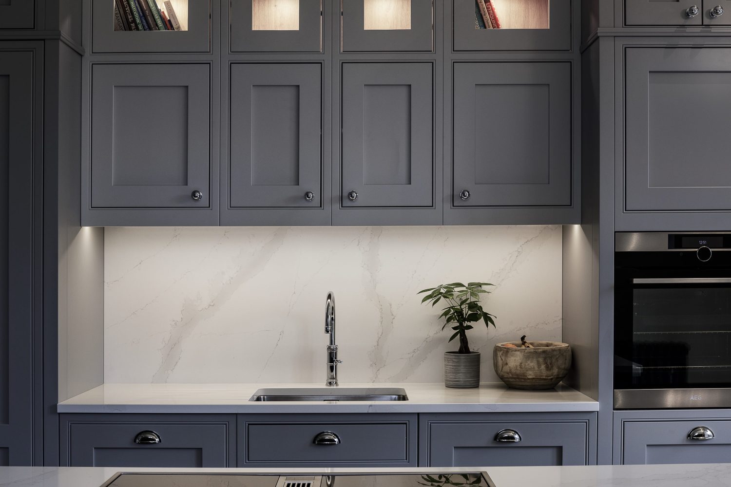 Grey kitchen cabinets with a white marble countertop and backsplash featuring a silver sink beside plant and fruit bowl