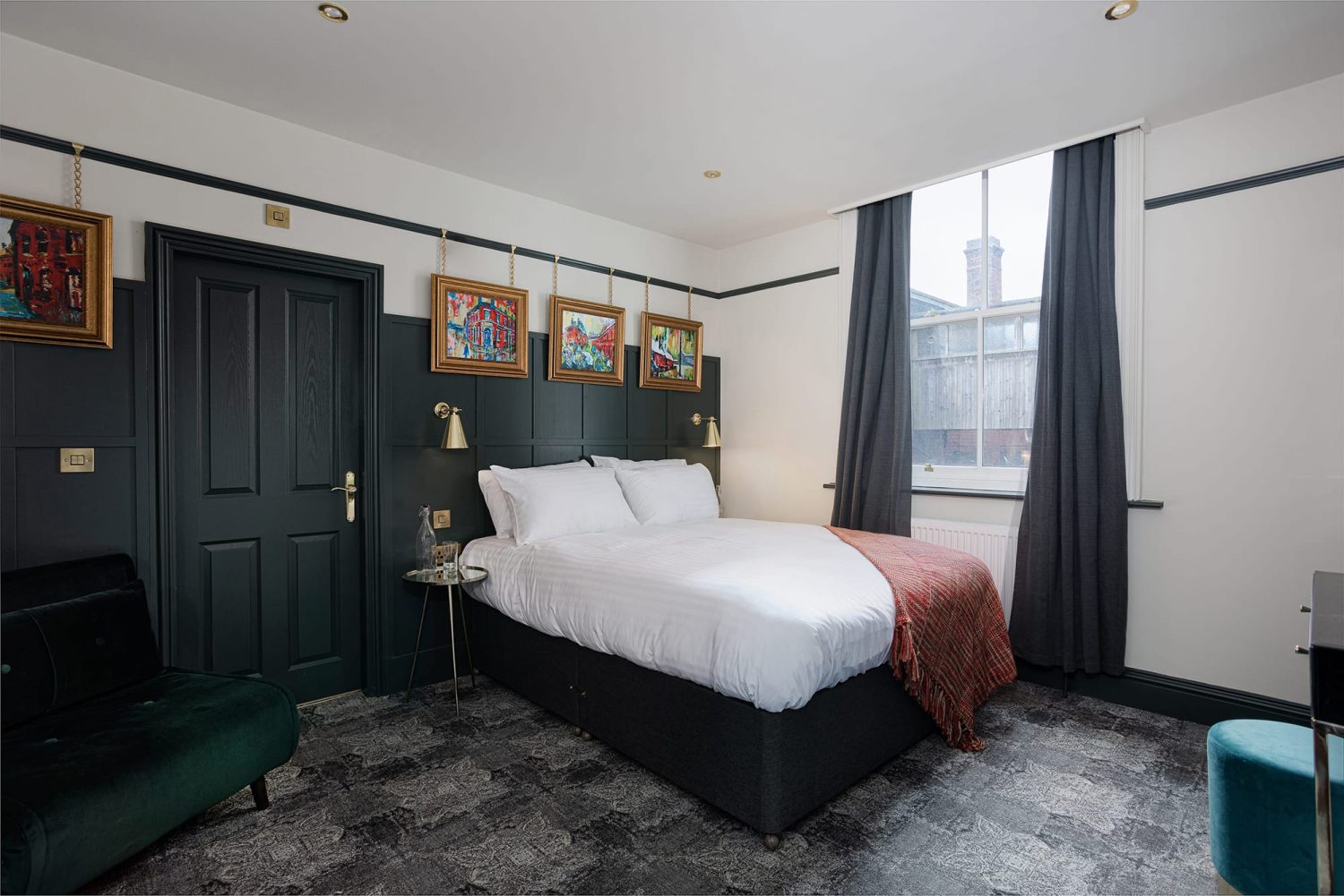 Double bed with dark panelled wall behind and gold framed artwork hanging on wall above and green velvet chair in corner