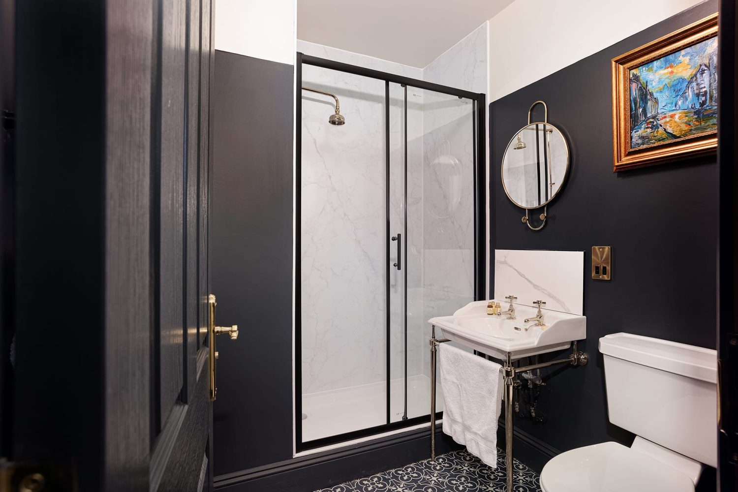 Bathroom with white marble tiled shower and dark painted walls with sink and toilet and gold mirror on wall