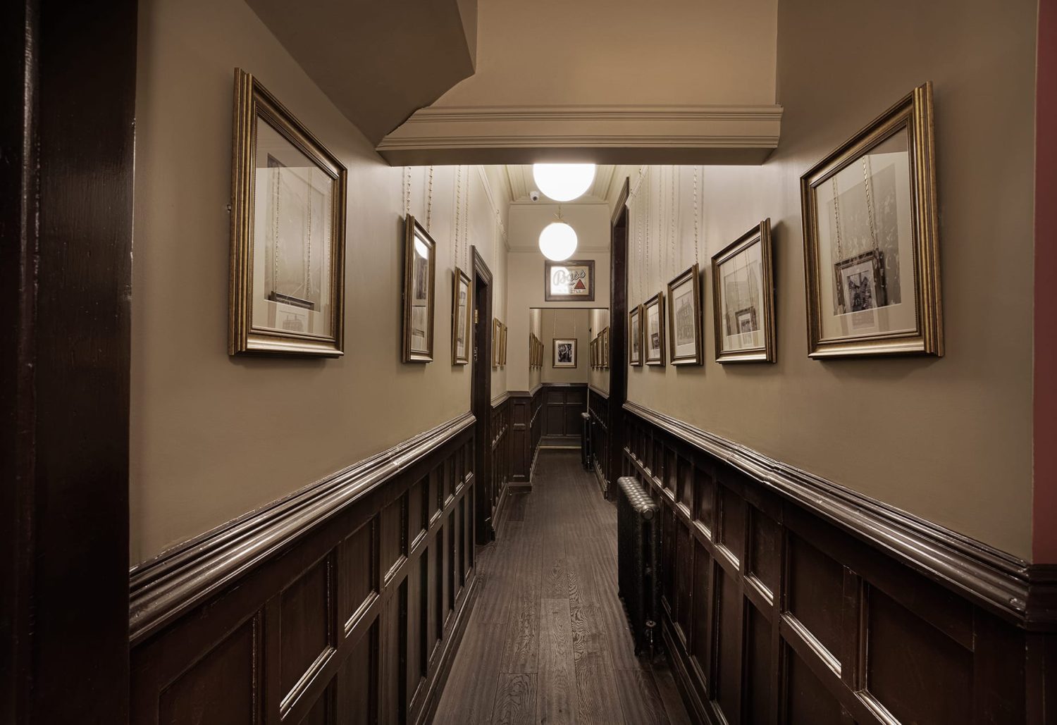 View down hallway with dark brown panelled walls and gold framed photos along walls and sphere pendant lights hanging from ceiling