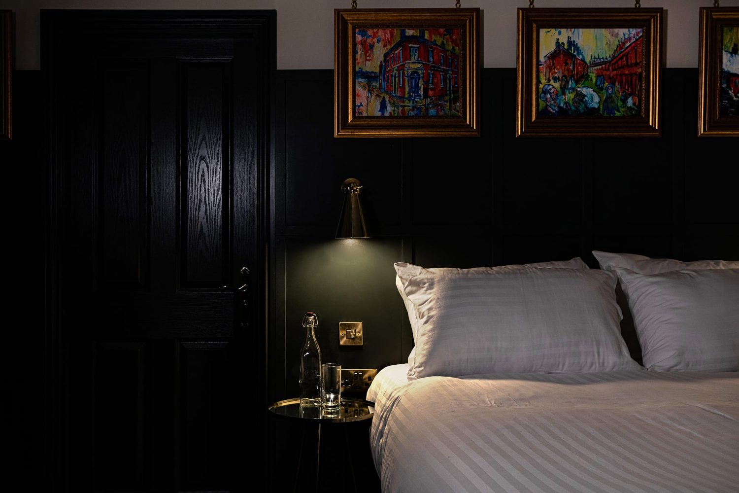 Corner of double bed with wall lamp glowing above bedside table and gold framed artwork hanging above bed