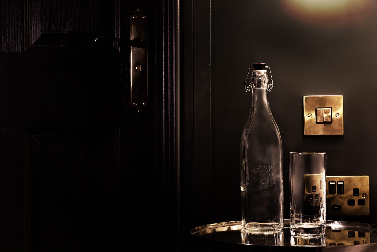 Small mirrored circular table with bottle and glass on top with gold light switch above on dark painted wall