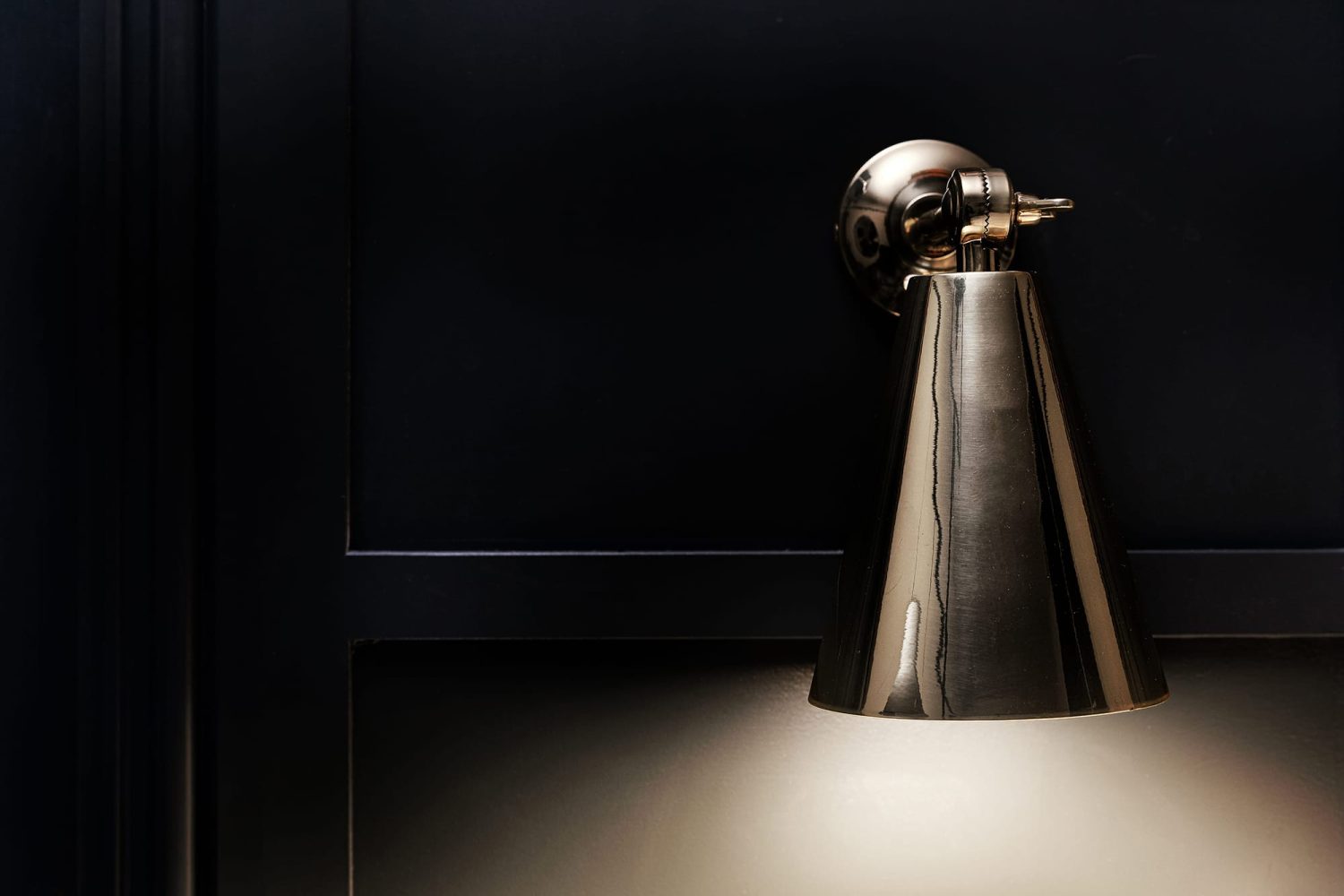 Silver cone shaped wall lamp glowing on dark panelled wall