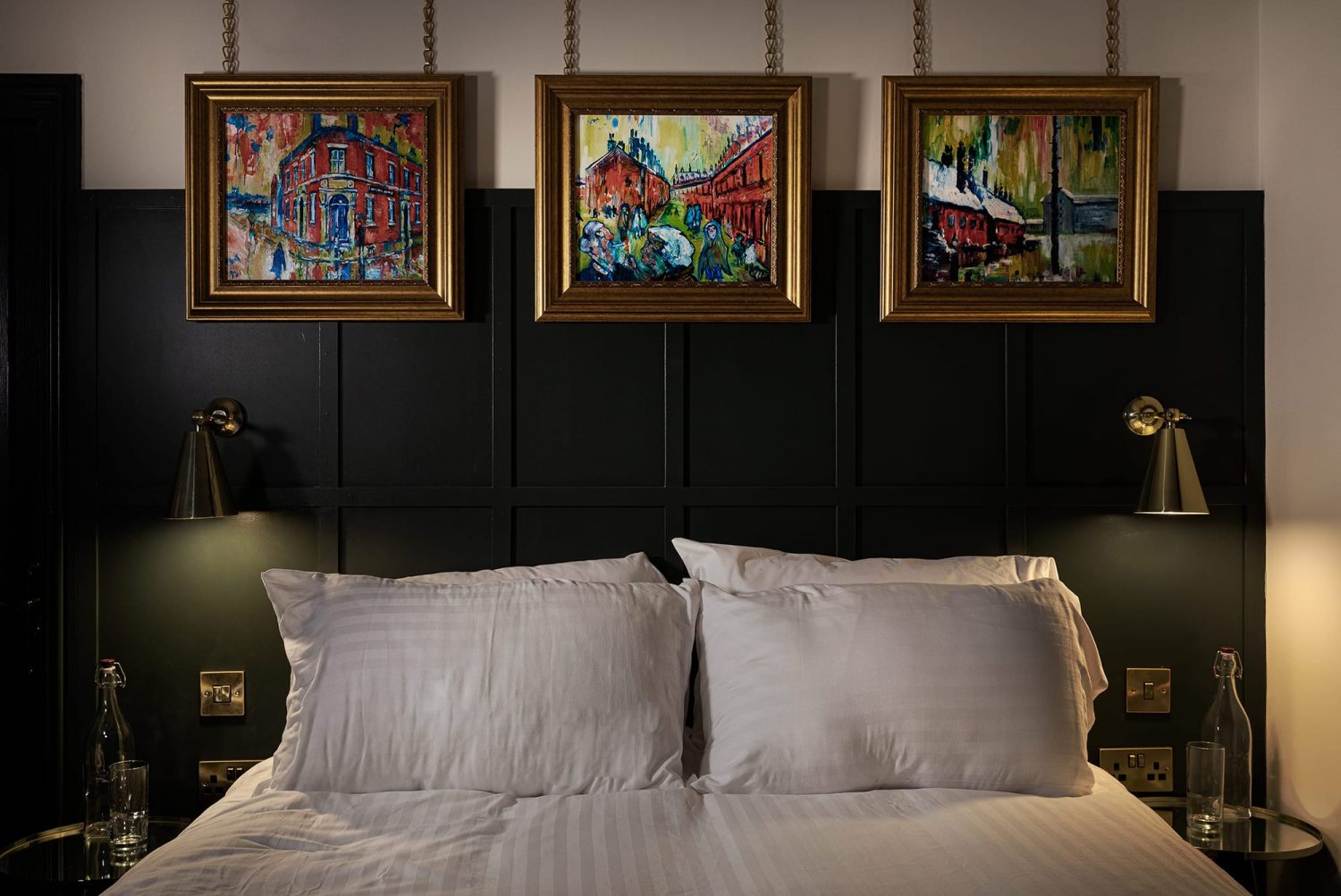 Double bed with dark panelled wall behind with gold framed paintings hanging above and cone shaped wall lamps to the sides