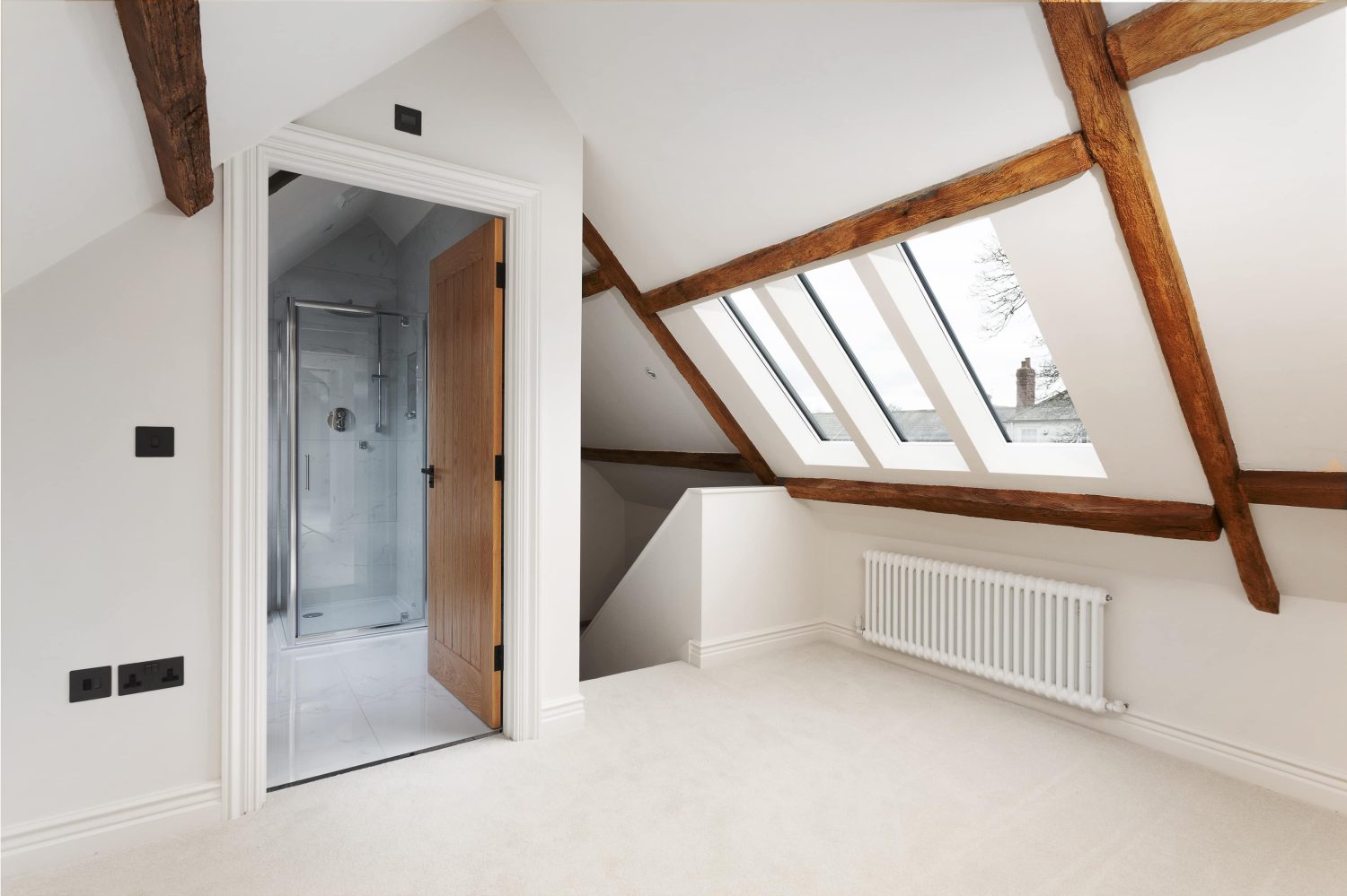Room with white walls and slanted ceiling with wooden beams looking into bathroom through doorway