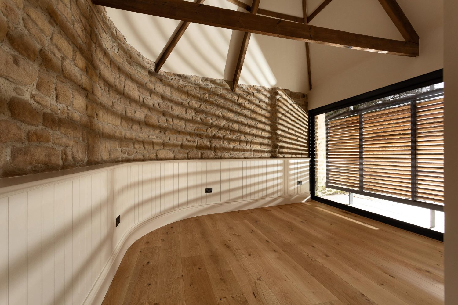 Curved wall of exposed stone and white wood across from wooden shutters over sliding door with sun shining through