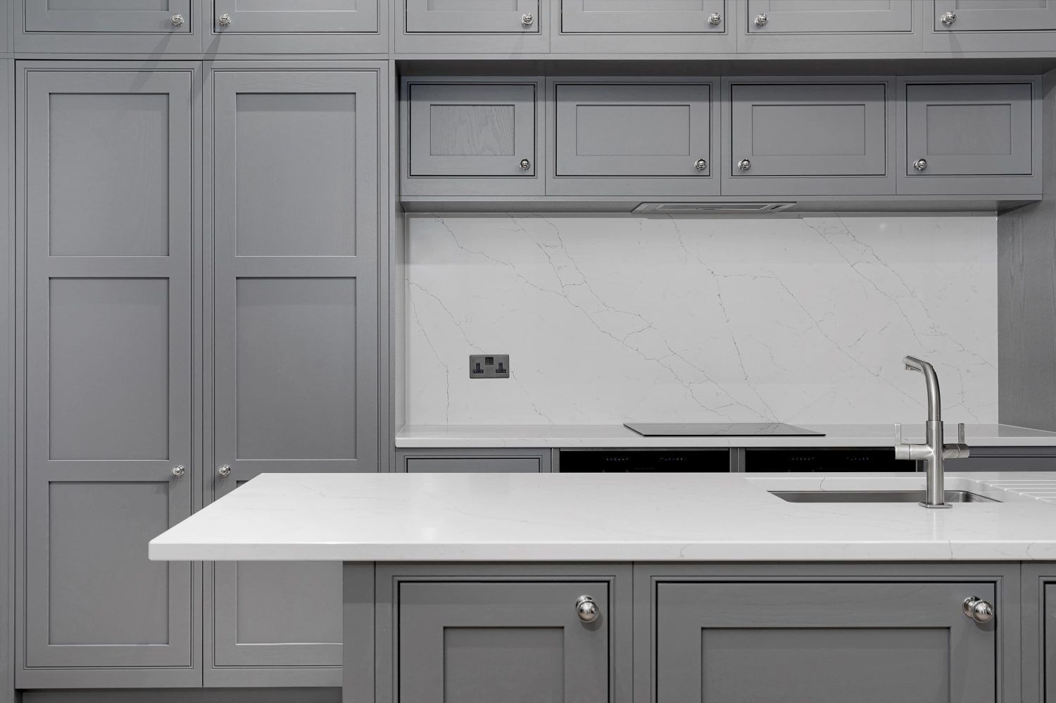 Kitchen island with silver tap in front of grey kitchen cabinets and white marble stove area