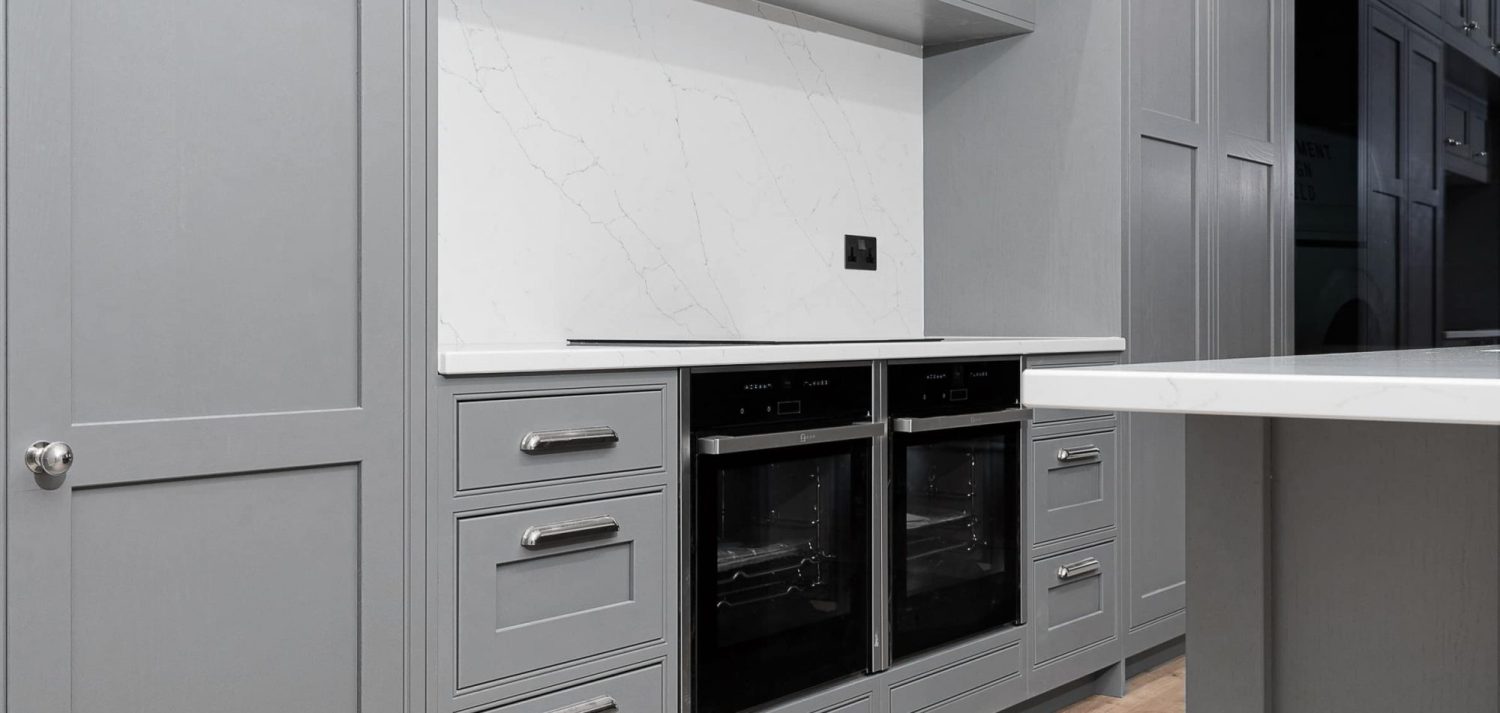 Double oven and stove unit surrounded by grey kitchen cabinetry and white marble back splash