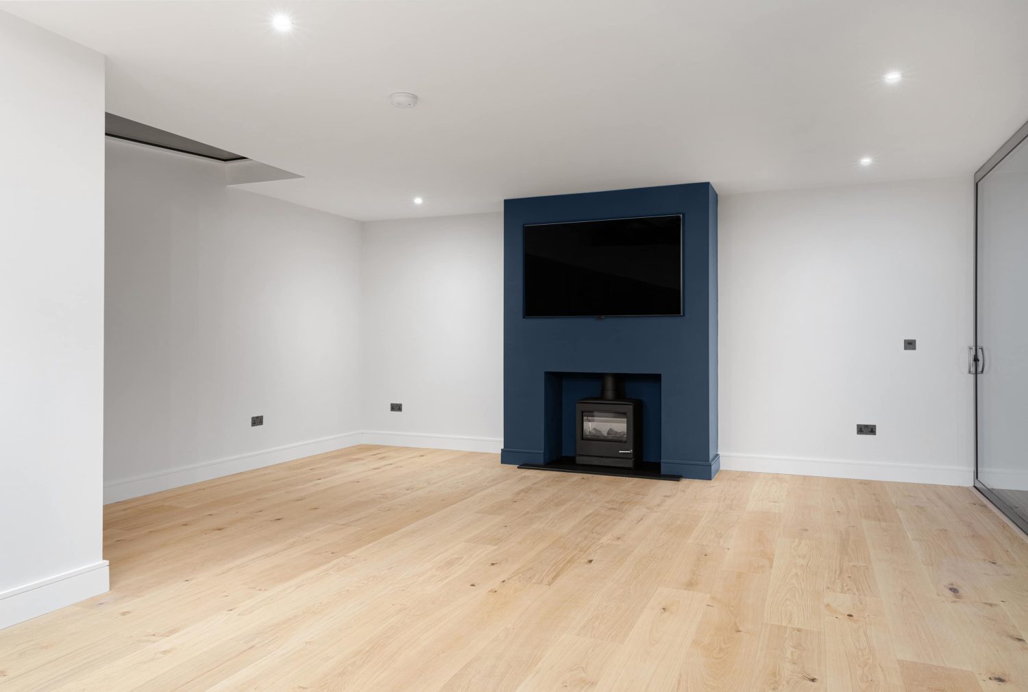 Room with white walls and wooden floor and central dark blue media wall with large TV above log burner