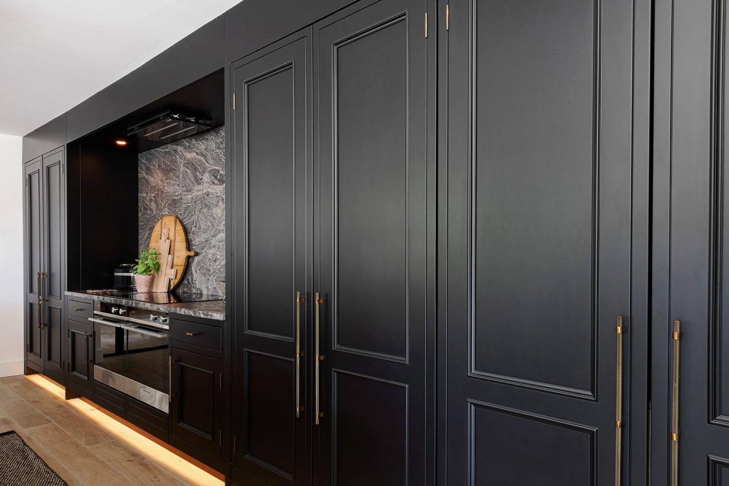 Black kitchen cabinets across wall with tall double doors with long gold handles and a dark grey marble back splash behind stove