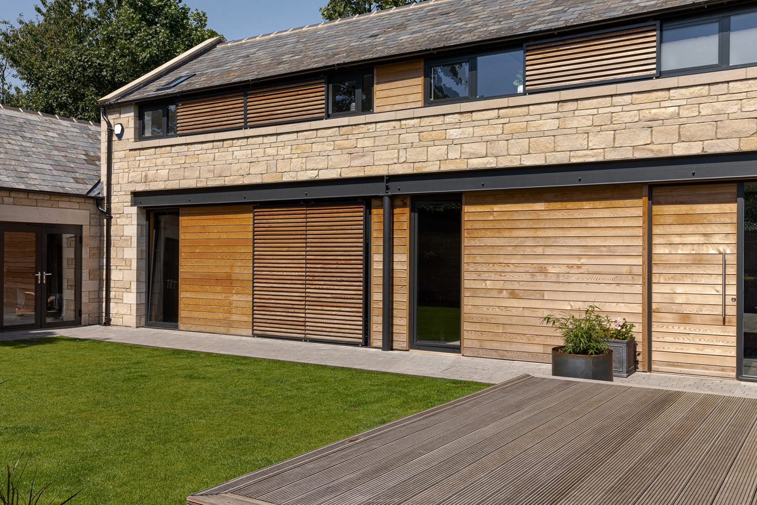 Exterior of stone barn conversion with wooden shutters over windows and wooden decking beside grass area