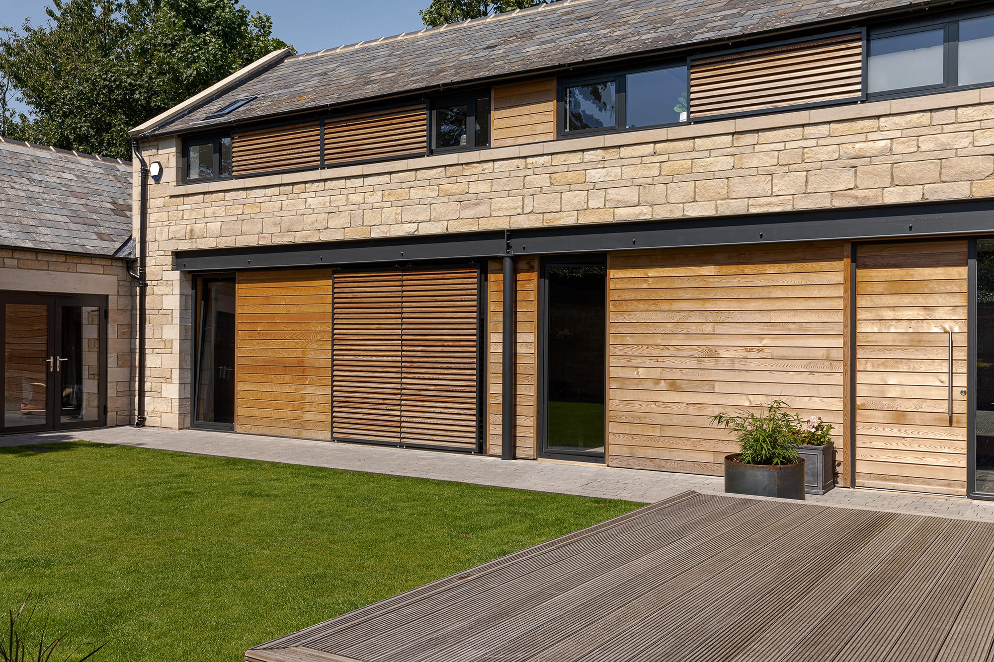 Exterior of stone barn conversion with wooden shutters over windows and wooden decking beside grass area
