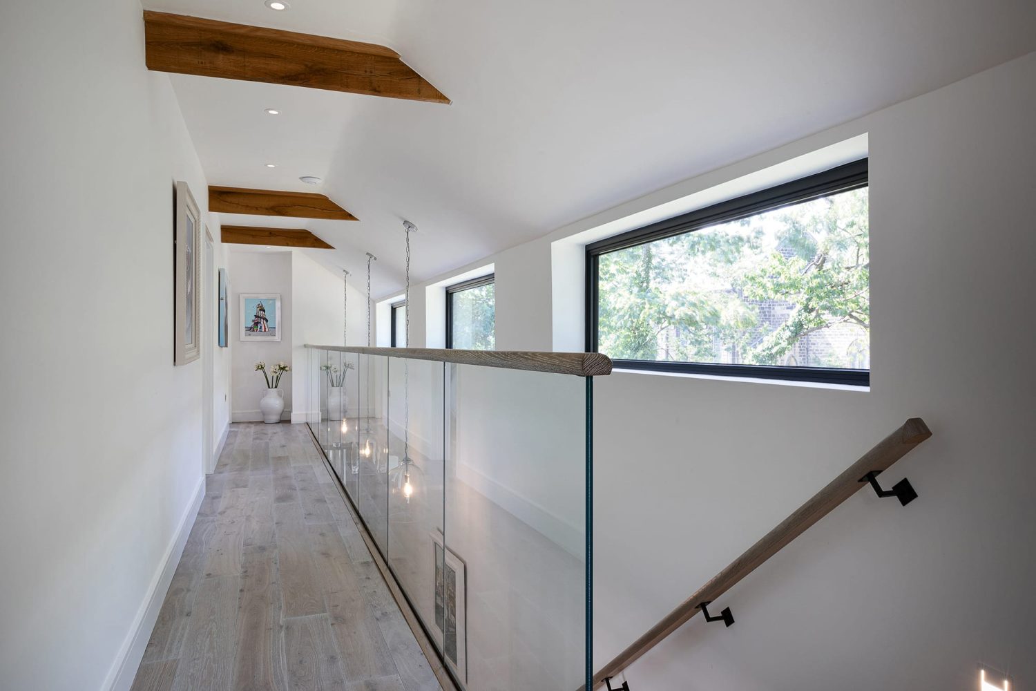 Upper floor landing with white walls and wooden flooring and glass stair barriers beside horizontal windows