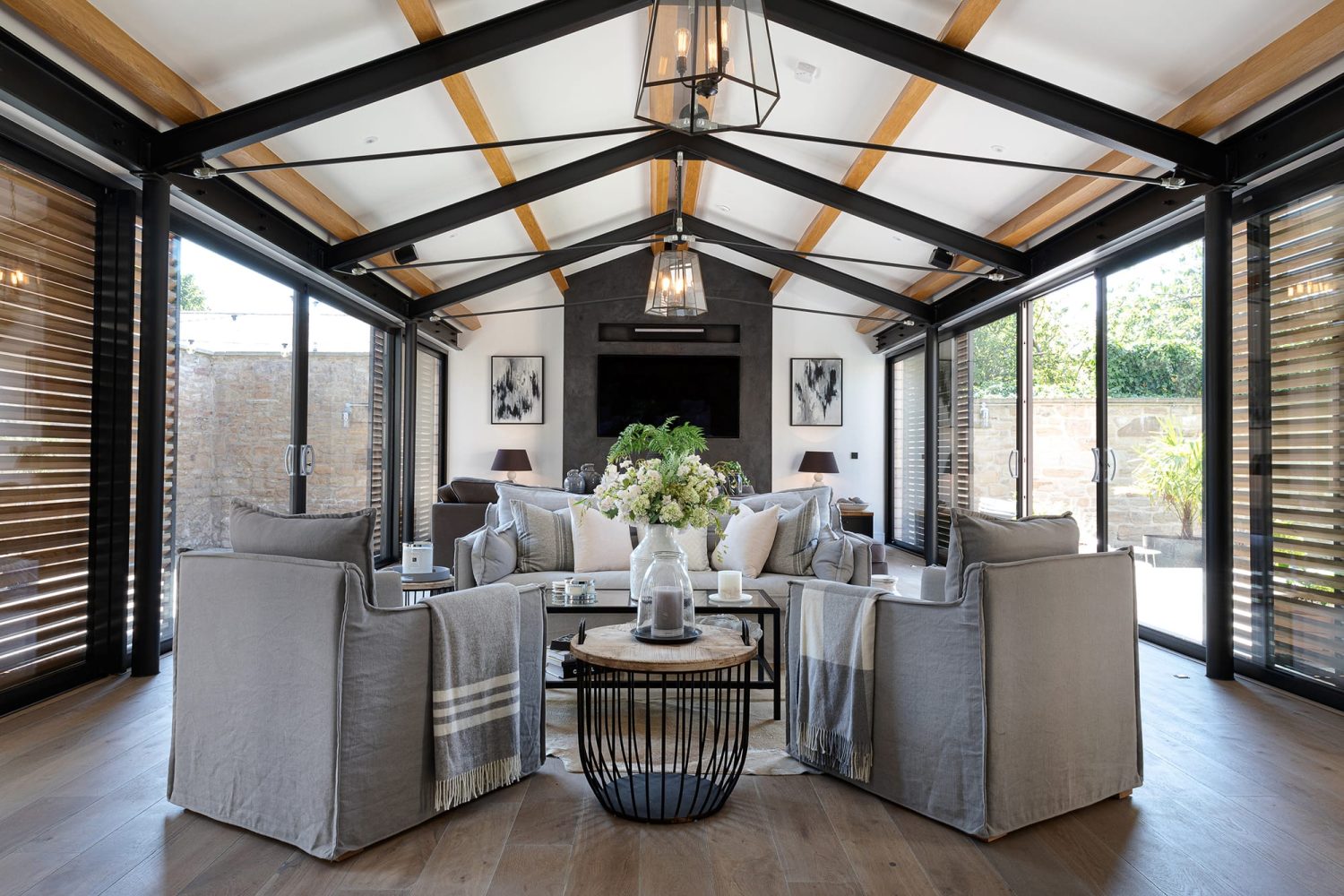 Living room with large glass sliding doors and beams on ceiling above grey sofa and chairs with large flowers as centrepiece