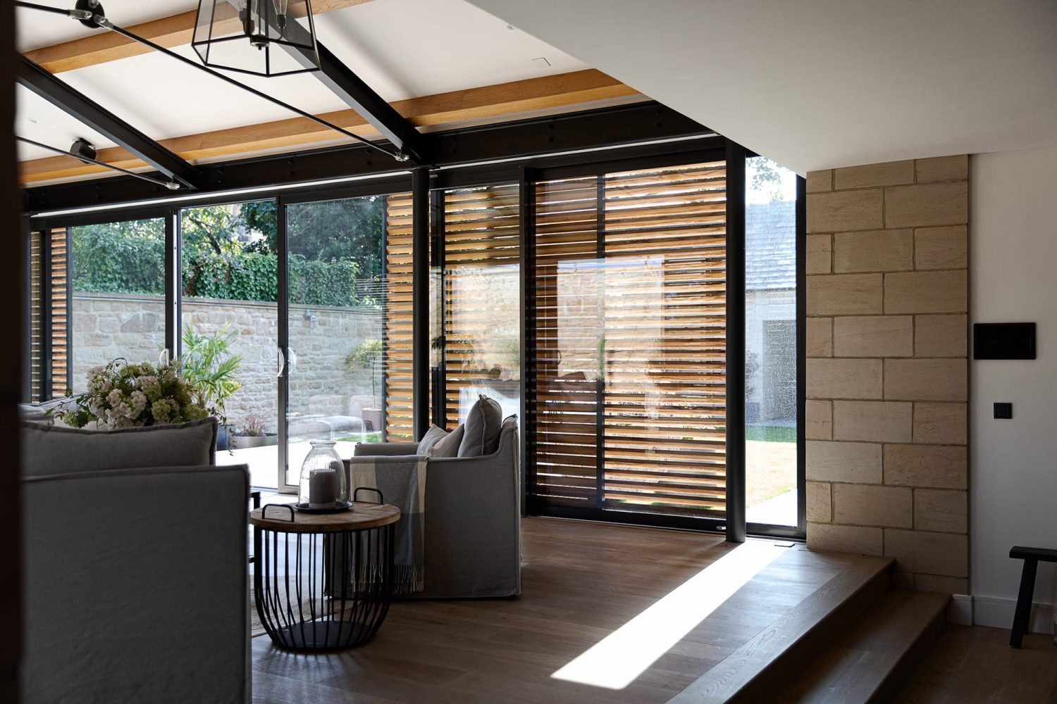Corner of living room with grey sofas and exposed stonework beside large windows with black frames and wooden shutters