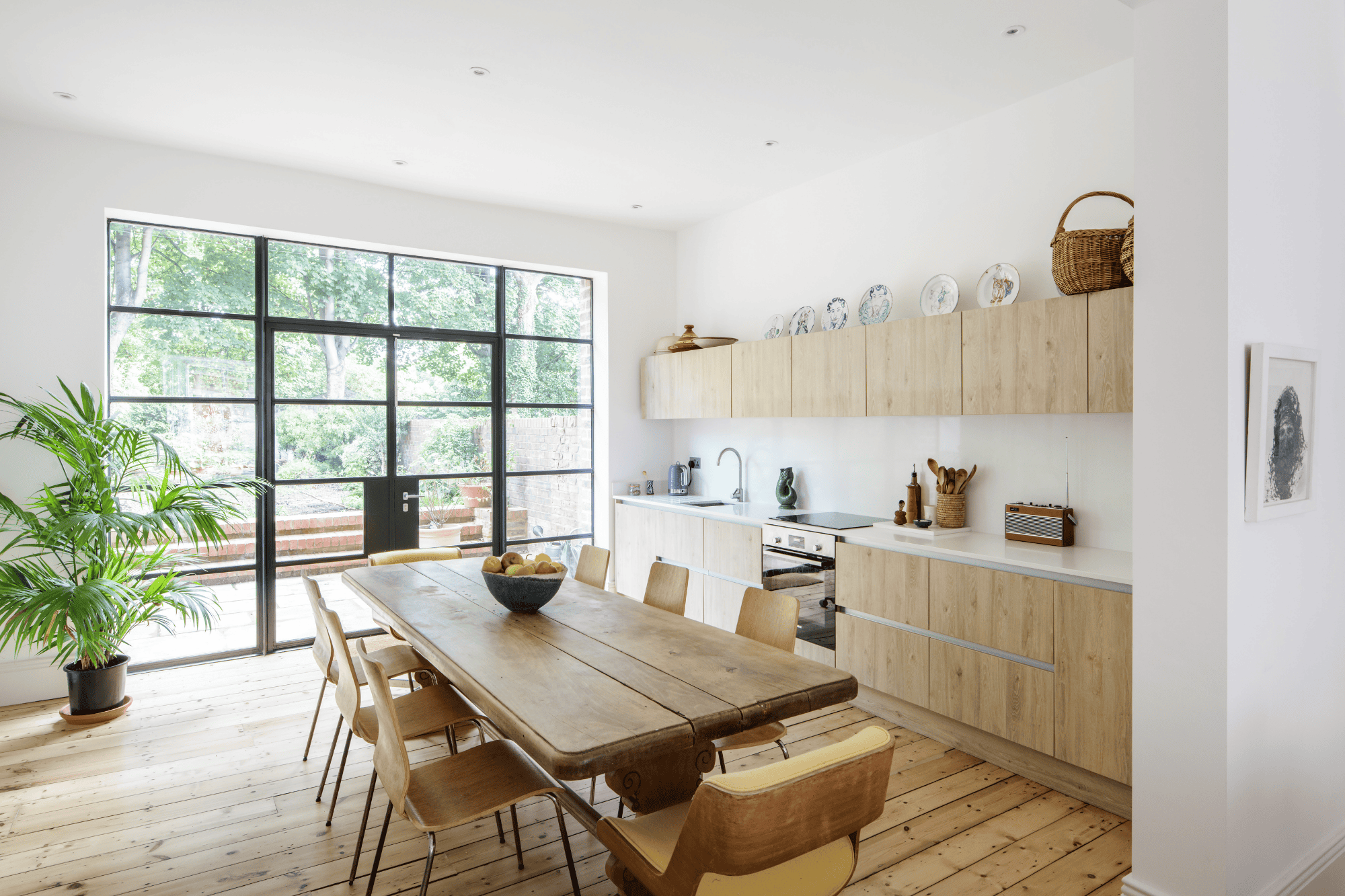 Kitchen with wooden cabinets and wooden dining table in the centre. Crittall style double doors leading outside with large leafy plant to the left.