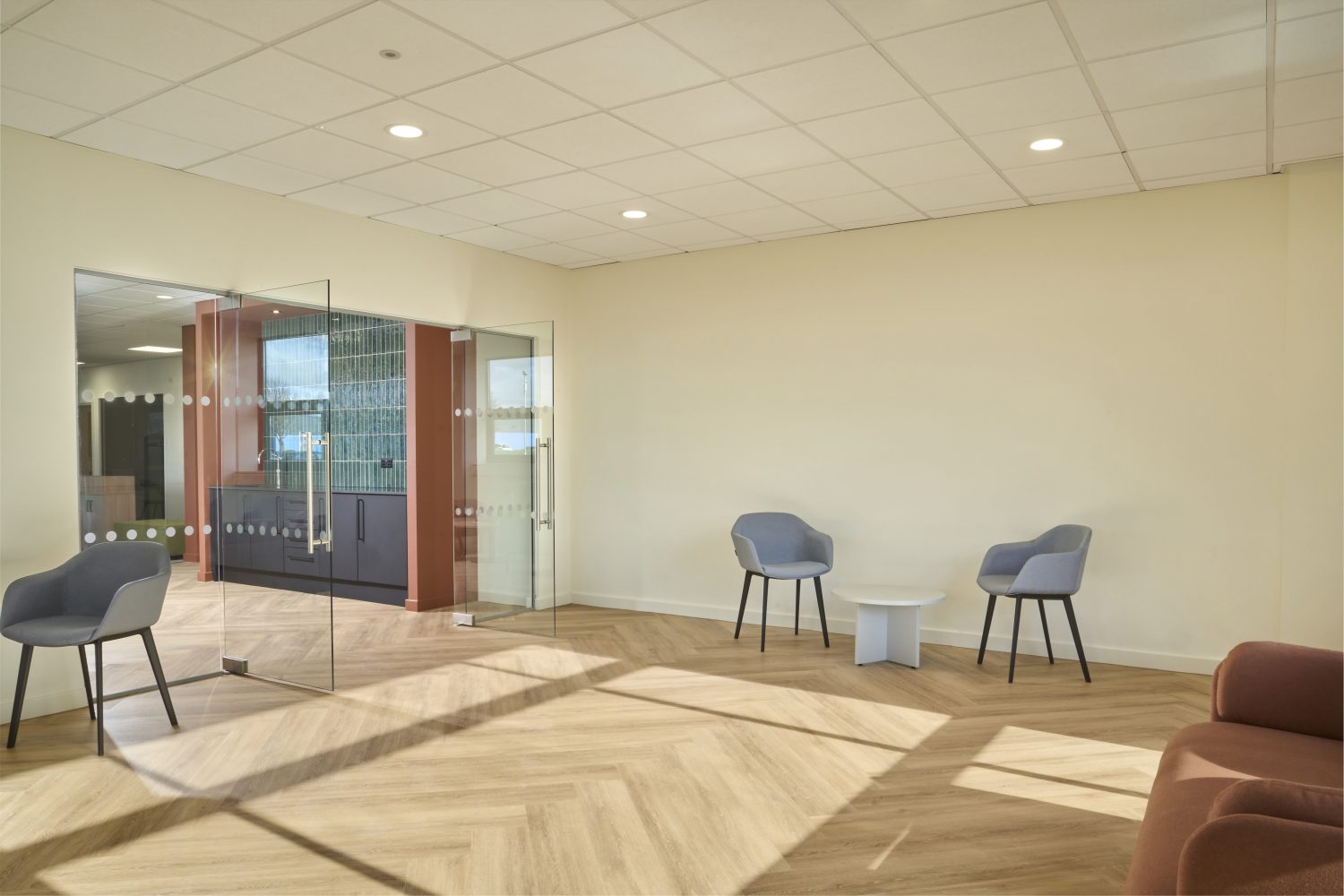 Entrance to offices with white walls and wooden floor. Corner of a muted orange sofa to the right with small white circular tables and grey chairs beside. Large glass doors opening into office