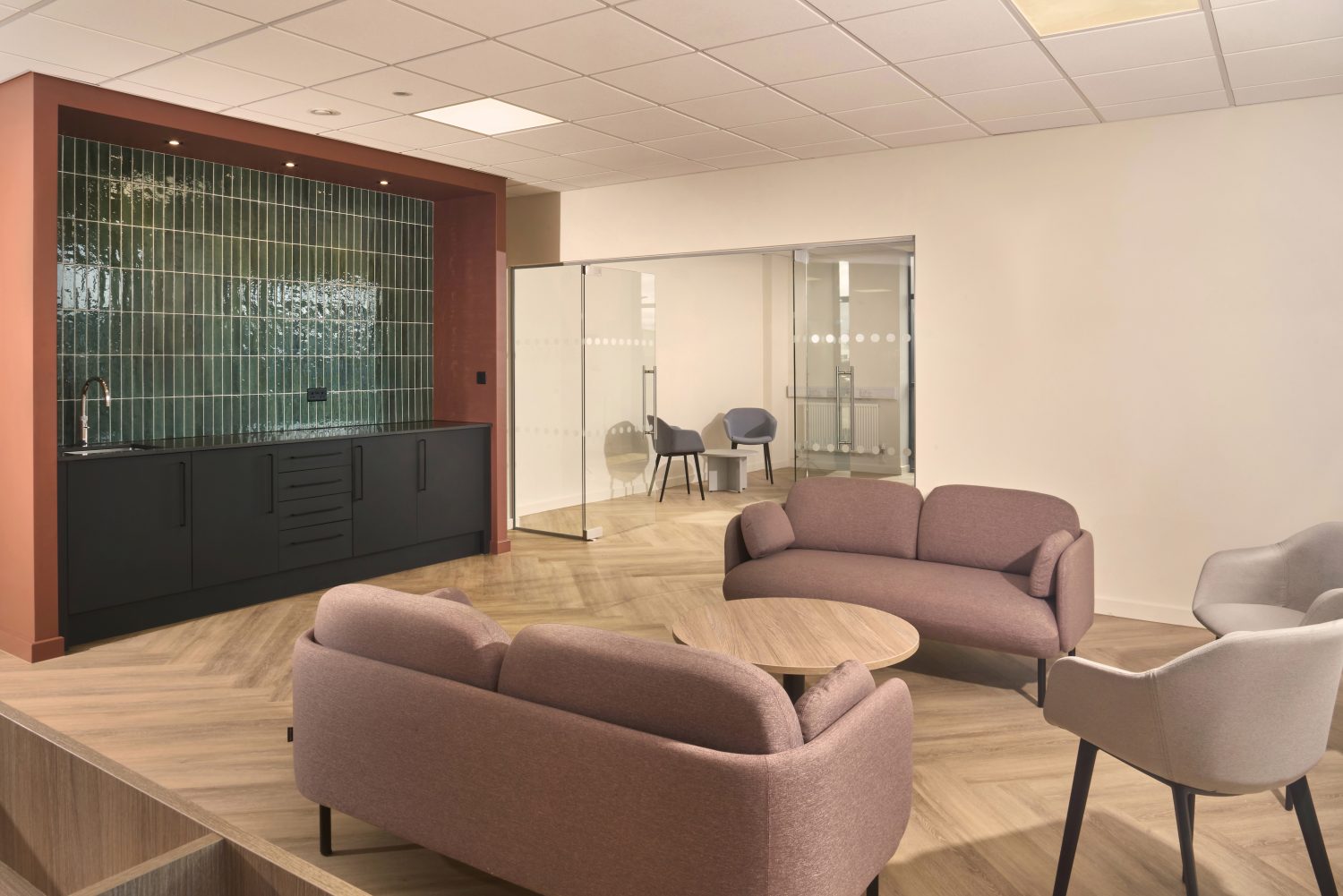 Two sofas and two chairs surrounding a circular, wooden coffee table next to office tea point with muted orange walls surrounding a green tiled wall and black cupboards
