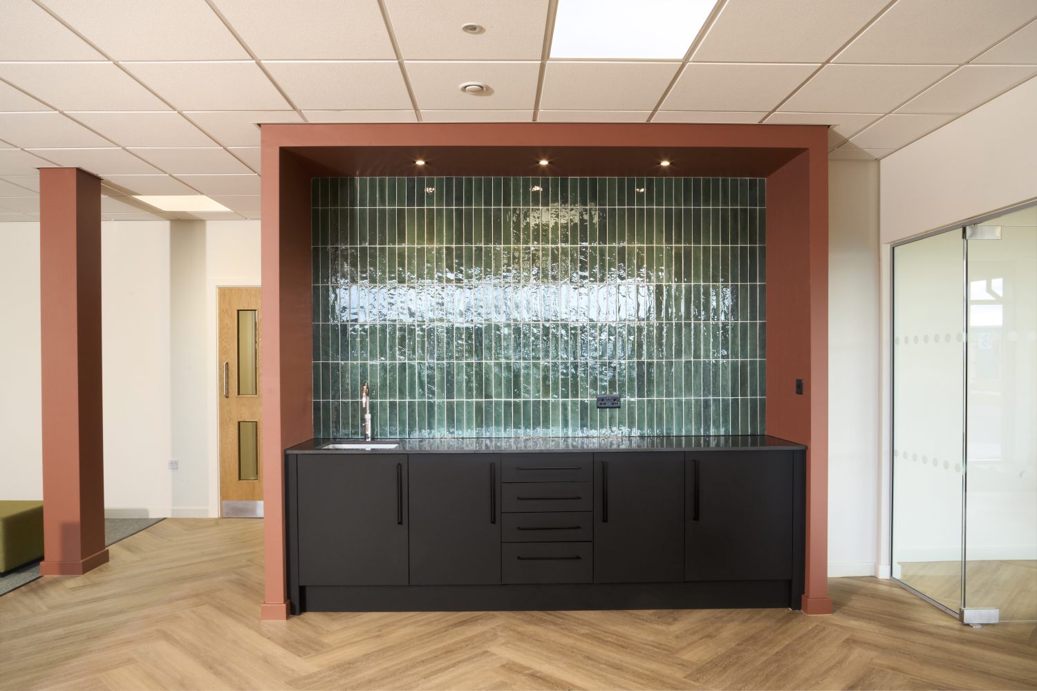 Office tea point with muted orange walls surrounding a green tiled wall and black cupboards with a black countertop and silver sink