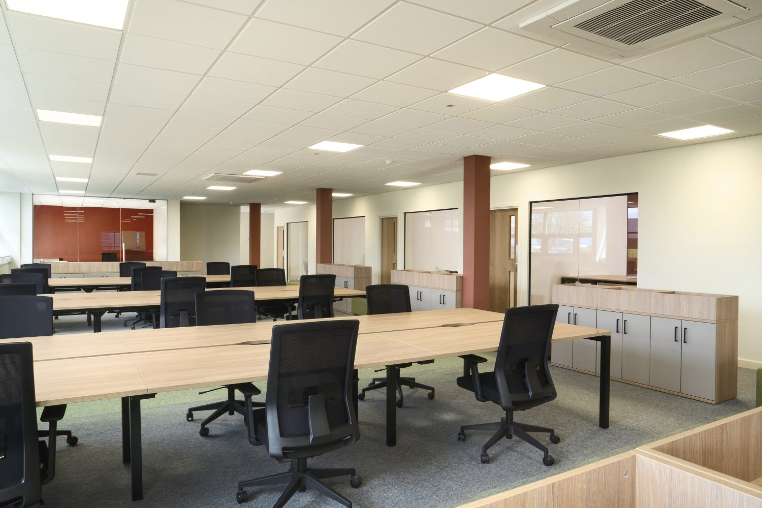 Open plan office with rows of wooden desks and black desk chairs beside wooden cupboards and glass doors looking through to offices