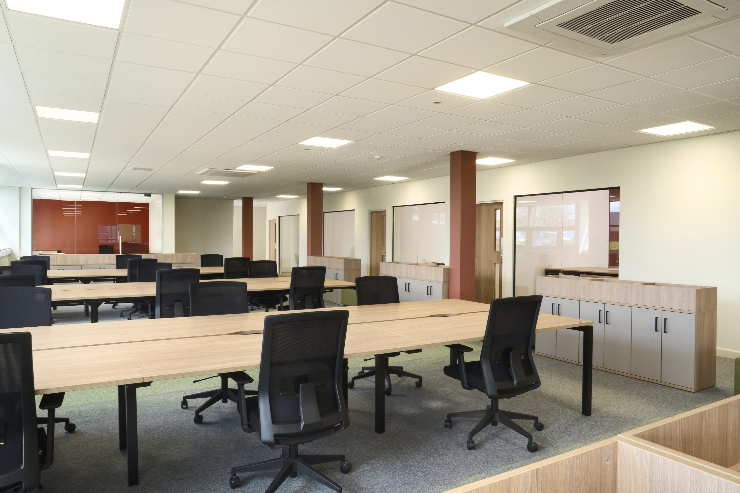 Open plan office with rows of wooden desks and black desk chairs beside wooden cupboards and glass doors looking through to offices