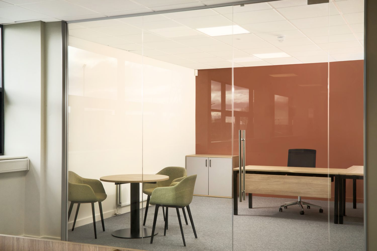 Single office with glass doors and muted orange wall. One wooden desk and a small, circular wooden table with three green chairs surrounding.
