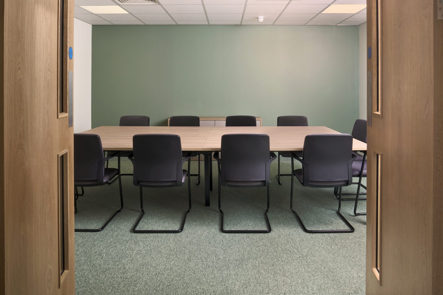 Meeting room with muted green wall and carpet and a large, rectangular table in the centre with black chairs surrounding.