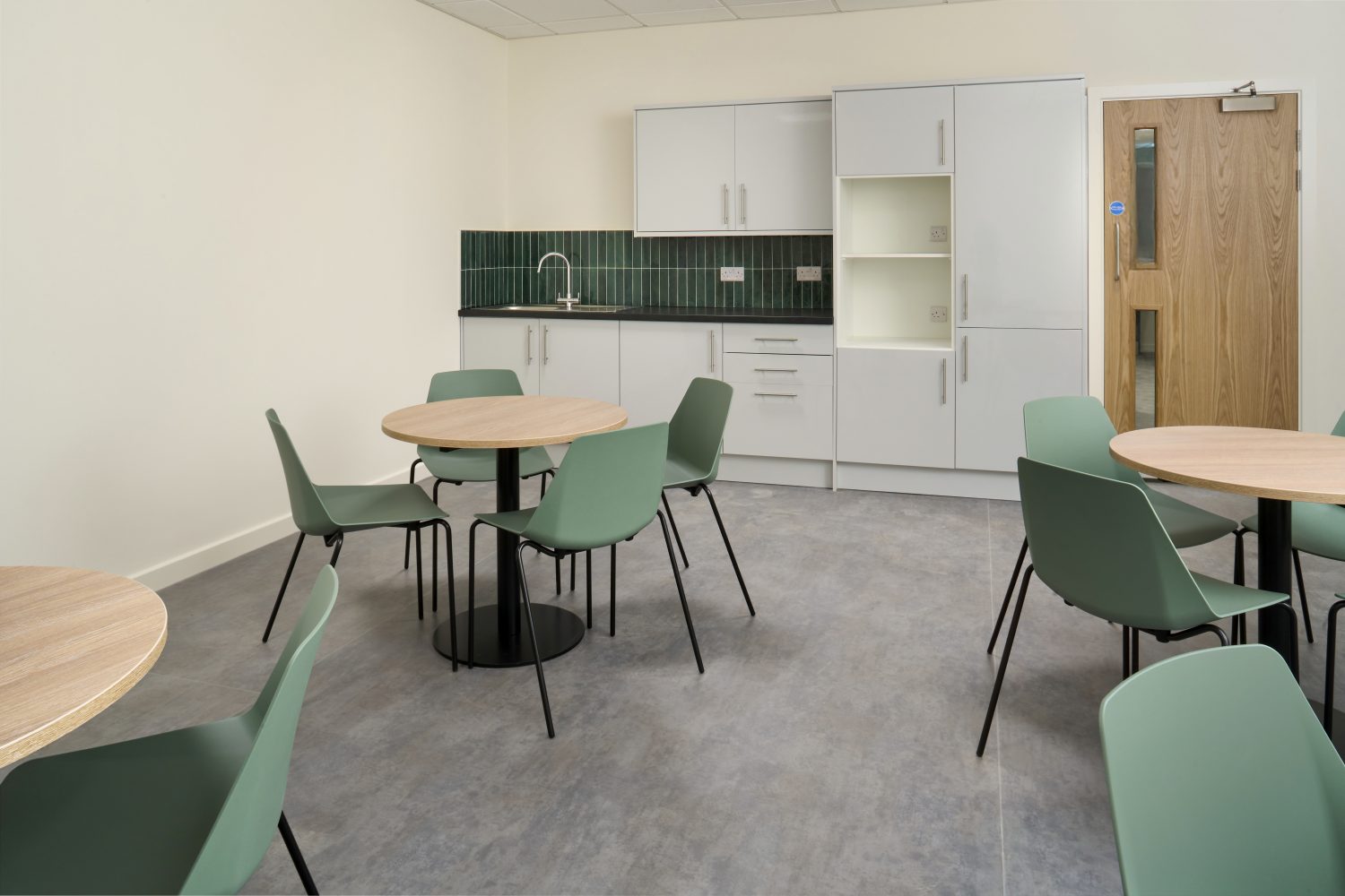 Small kitchen with white cabinets and black countertop with green tiled backsplash. Circular wooden tables with four green chairs each.