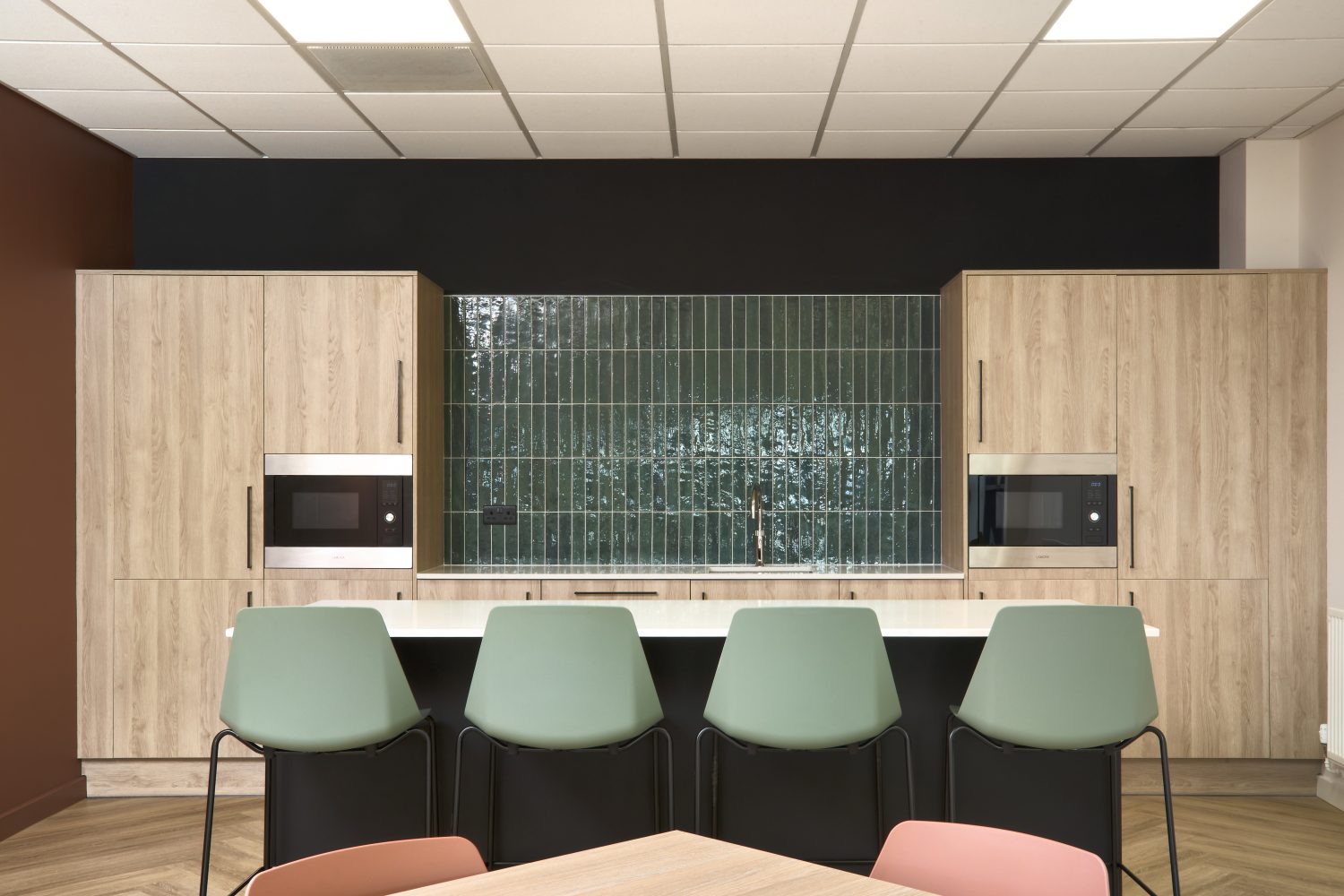 Kitchen with wooden cupboards and green tiled backsplash in the centre with an island in front and muted green chairs