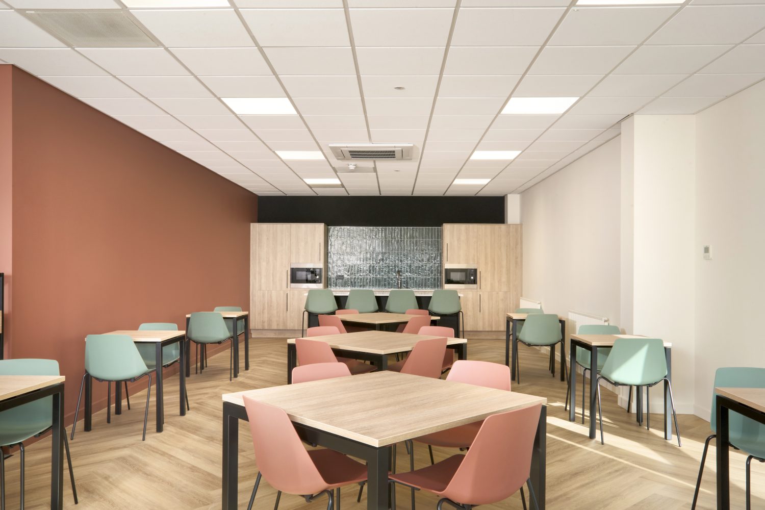 Kitchen with muted orange and white walls and wooden cupboards on the far wall with green tiled backsplash. Wooden tables with muted orange and green chairs