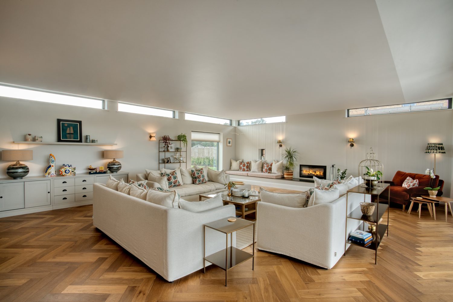 Overview of living room with three white sofas on wooden floor and cream walls with thin horizontal windows