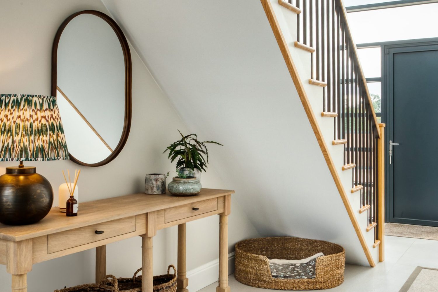 Hallway with wooden table beneath staircase with oval wall mirror and wicker baskets
