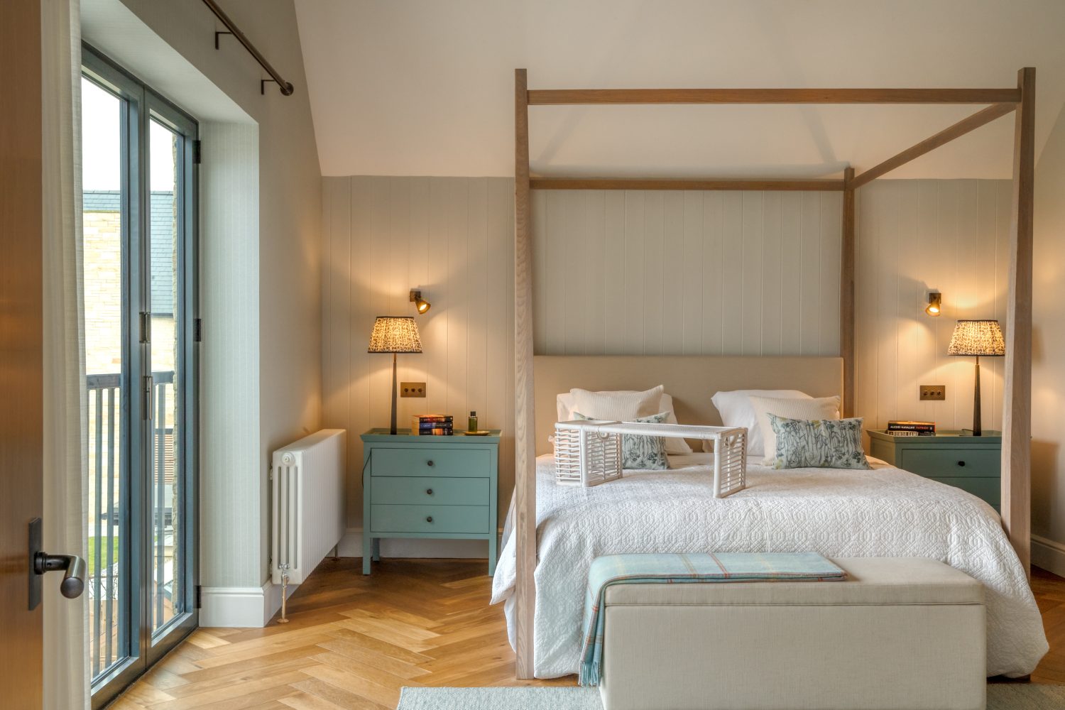 Wooden four poster bed with muted green beside drawers on either side with cream panelled wall behind and juliette balcony to the left