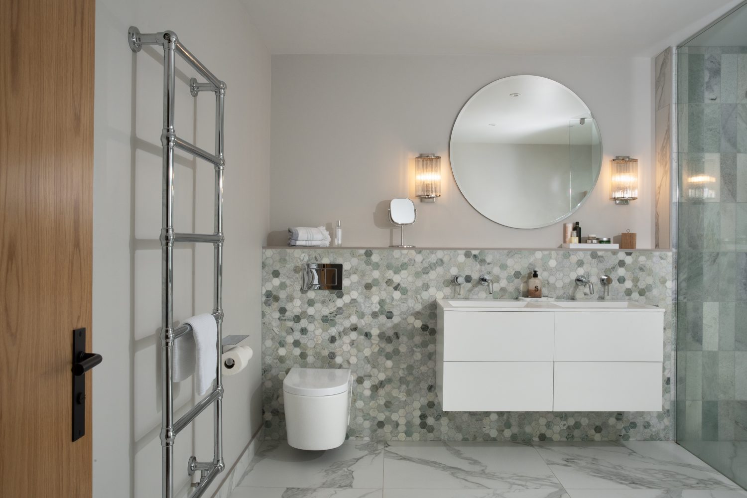 Bathroom with green tiled wall behind white toilet and double sink unit with circular mirror and lamps on wall above and silver towel rail to the left